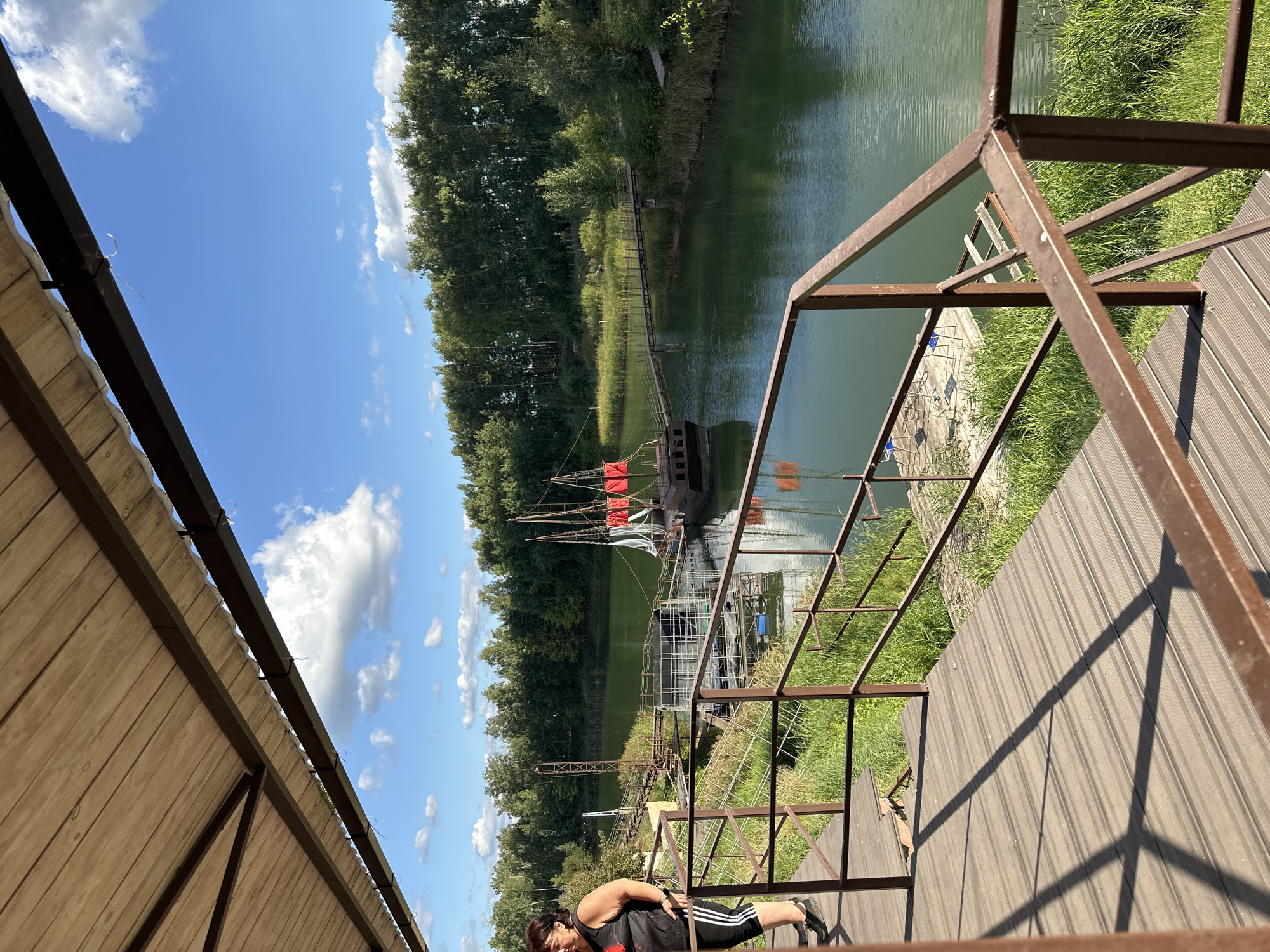 An overhead view of a wooden deck area with a woman standing on it, looking out at a body of water. The deck has a wooden planked surface and is bordered by a brown metal railing. Beyond the deck, a calm green body of water is visible, with a wooden bridge crossing it and a boat with red sails docked in the water. The background is a dense line of green trees under a bright blue sky with scattered white clouds. The overall scene suggests an outdoor recreational area, possibly a park or waterfront attraction, located in the village of Пады, Russia.