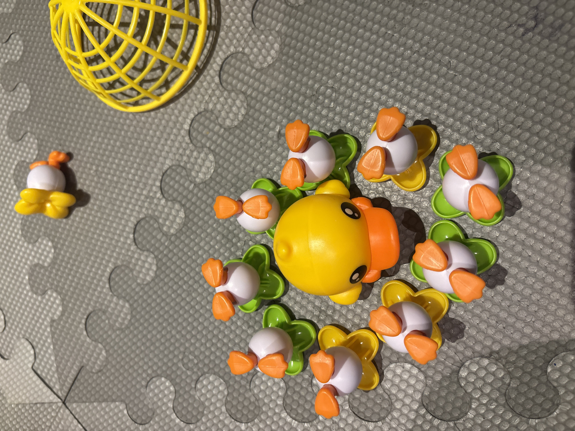 A close-up, high-angle shot shows a group of yellow and white plastic duck toys scattered on a grey, interlocking foam mat. A large yellow duck with orange beak and black eyes is positioned in the center, surrounded by several smaller white ducks with orange beaks and feet, each sitting on a colorful plastic flower-shaped base. In the upper right corner, a yellow plastic cage-like structure is visible, and in the upper center, a single yellow duck toy is lying on its side. The lighting suggests an indoor environment, and the toys appear to be arranged on the mat in a playful, somewhat random manner. The scene is devoid of people and appears to be a static display of children's toys. There is no information to suggest the location is in Dubai, United Arab Emirates.