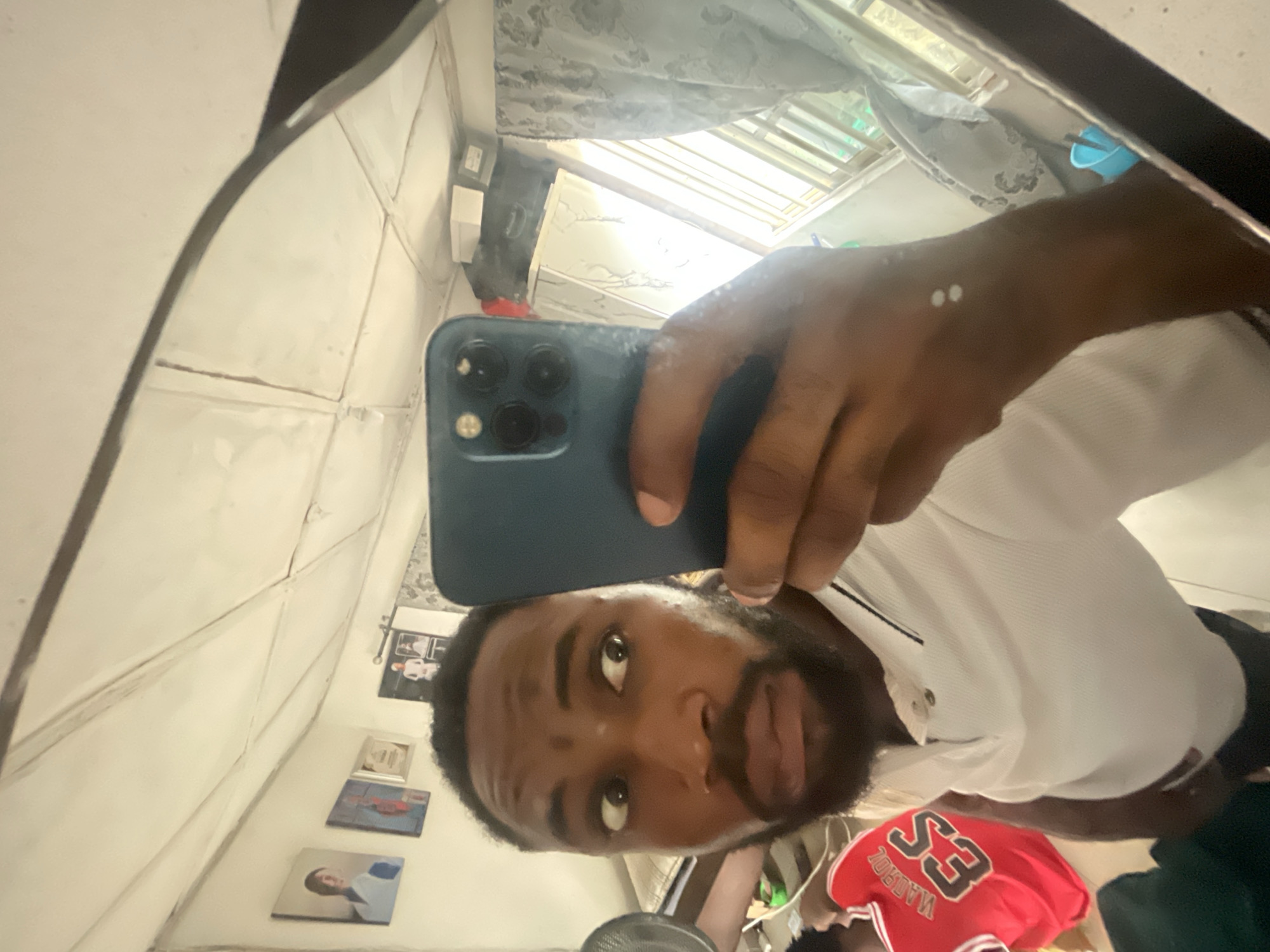 The image shows a selfie taken in a mirror. A man is holding a blue smartphone to capture the image. The background reveals a room with white walls decorated with framed images. Outside the window, the scene shows a view of the sky. The specific location in Lokoja, Nigeria cannot be determined from the provided image.