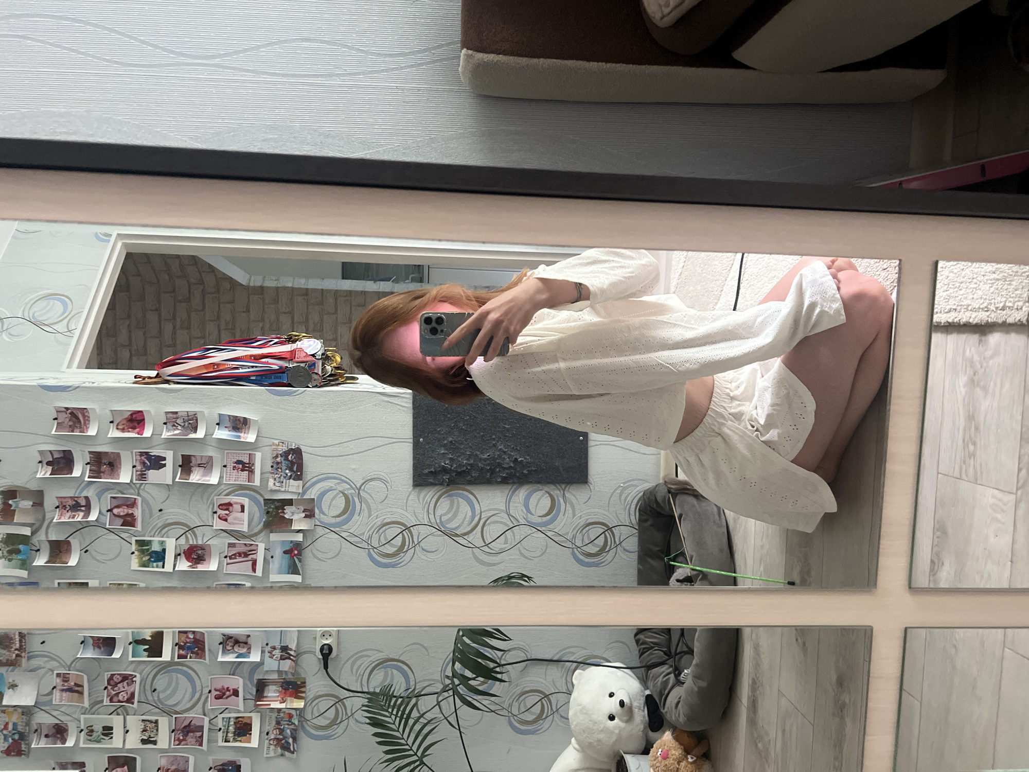 A person with long, auburn hair is reflected in a grid-patterned mirror. The person holds a smartphone to take a photo or video, dressed in a white, eyelet-patterned top and matching shorts. The reflection reveals a room decorated with a patterned wallpaper adorned with numerous small photographs. Below the wallpaper, there is a plant with green, fern-like leaves and various objects, including a white teddy bear. The setting could be a residential space, and the information suggests the possible location to be Chaikovsky, Russia.