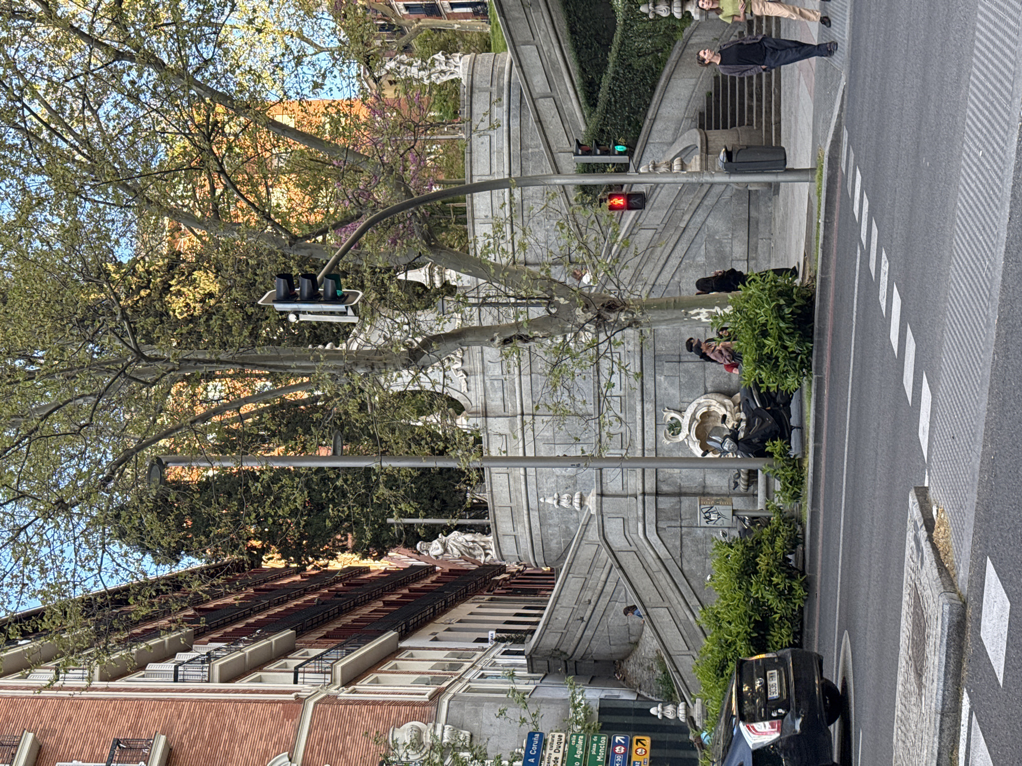 The image depicts a street scene in Madrid, Spain, notable for its urban environment and architectural features. A multi-story building with a red-tiled roof and rows of balconies dominates the left side of the frame. To its right, an imposing stone staircase leads upwards, adorned with classical sculptural elements, including a prominent statue. Pedestrians are present on the sidewalk alongside the staircase, and a few are positioned on or near the stairs themselves. A traffic light stands at the intersection with a red signal illuminated. A black car is parked at the curb. Street signs with route information are visible in the lower left corner. The scene is further embellished by trees with budding leaves that spread across the top of the image, partially obscuring the sky.