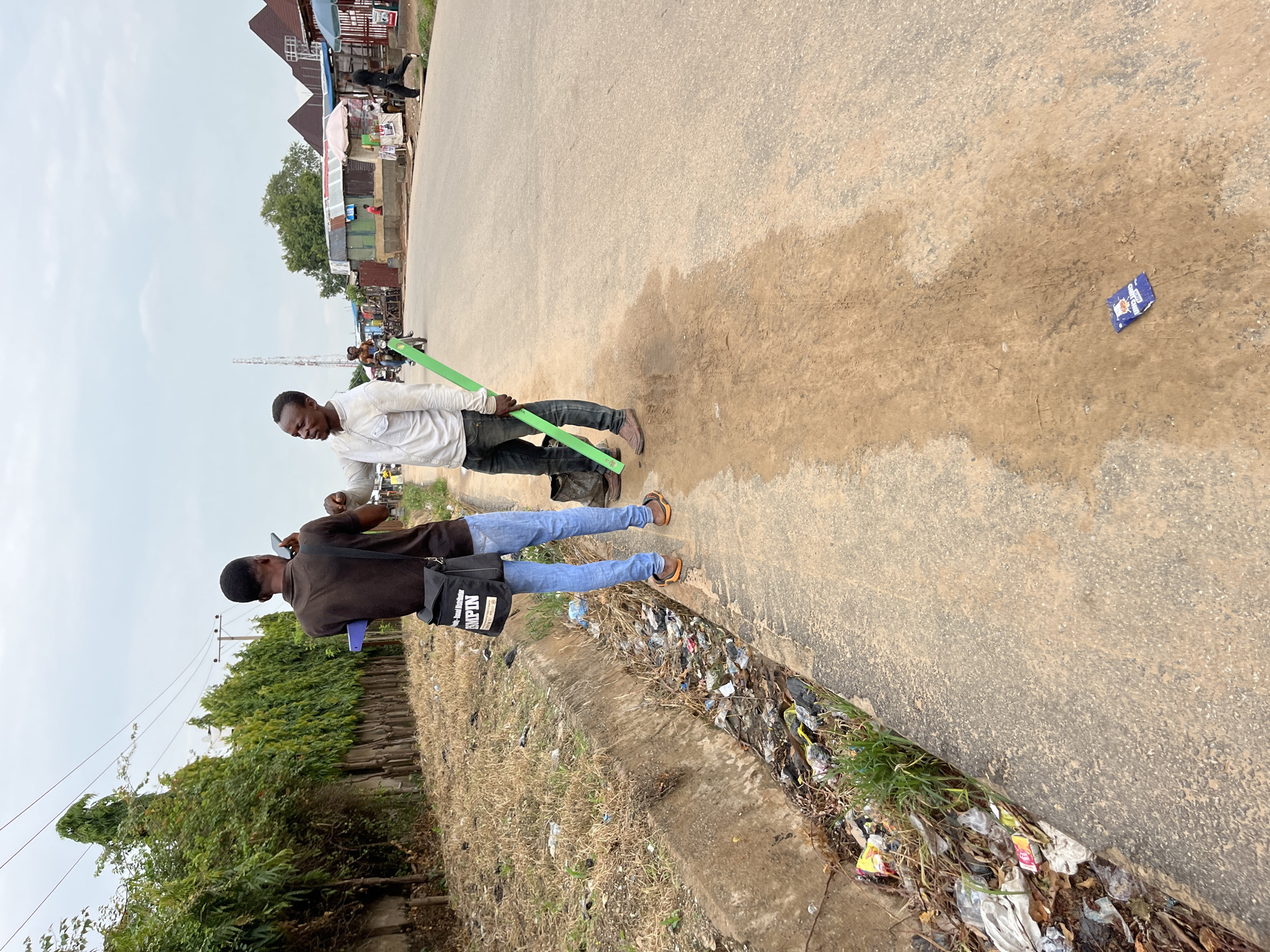 Two men stand on a paved road in Abuja, Nigeria. One man, wearing a white shirt and blue jeans, holds a long green object. The second man, wearing a brown shirt, blue jeans, and a black messenger bag, appears to be taking a picture or video with his phone. Debris lines the edge of the road where it meets vegetation. Buildings are visible in the background. The road appears to be slightly wet.
