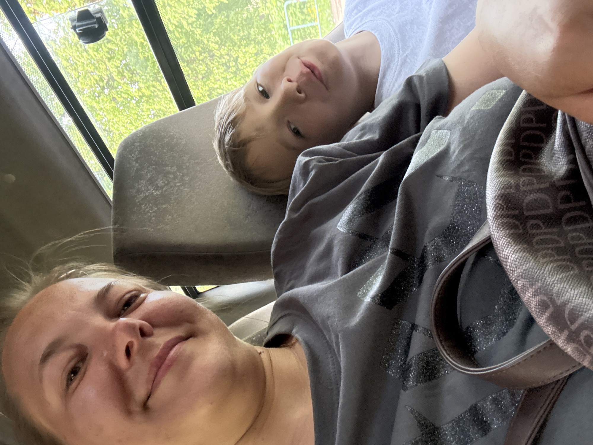 A woman and a young boy are inside a vehicle, possibly a van or bus, in Novosinkovo, Russia. The woman is in the foreground, partially out of focus, looking toward the camera. The boy is lying on the back of the seat. Both are wearing casual clothing. A patterned bag is partially visible. Outside the vehicle, there are trees.