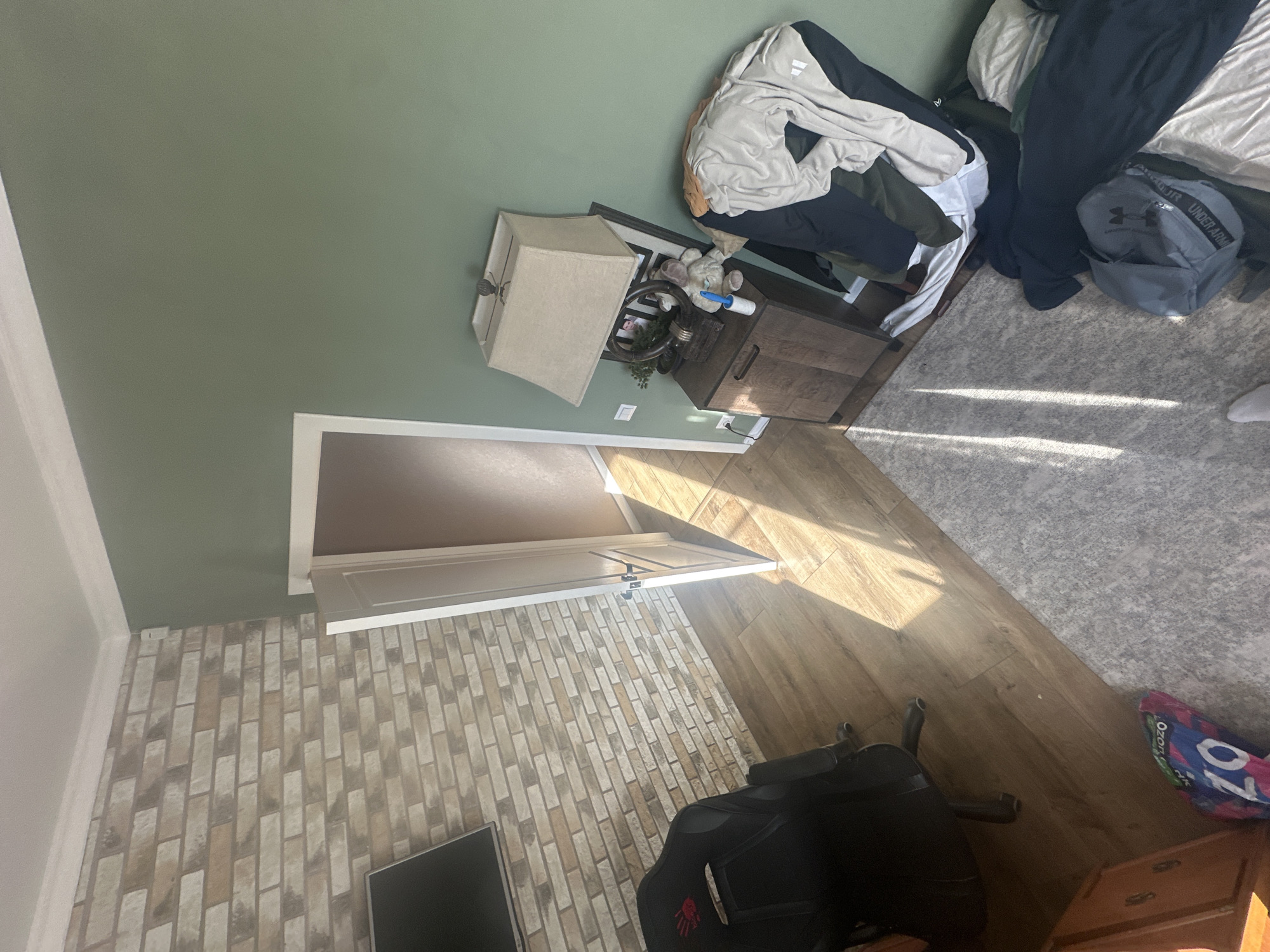 The image shows a room with green walls and a patterned brick wall, which could be a room in a house in an unknown city. The floor is made of wood panels. In the top right corner is a bed with clothes and an Under Armour backpack on it, next to a grey carpet. A nightstand with a lamp on it is near the bed. An open door leads to another room. In the bottom center of the image, there is a black gaming chair near a TV screen and a wooden chest.