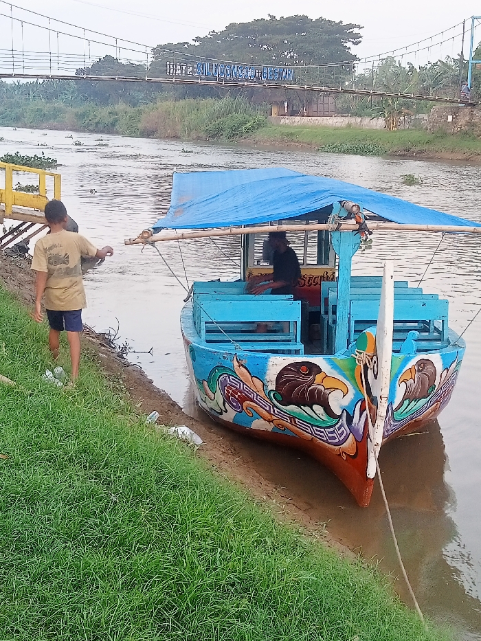 The image shows a brightly colored boat, docked at the bank of a river in Jakenan, Indonesia. The boat has a blue canopy, blue seats, and a brightly colored painted hull with stylized, eagle-like motifs. It is moored along the bank of the river. One man, wearing a blue shirt and khaki pants, is seated in the boat, while another man wearing a brown t-shirt and khaki shorts, is standing on the bank beside the boat. The boat is near a suspension bridge that crosses the river. The bridge is adorned with a sign with the words "Jembatan Gantung Bestari". The river is calm and surrounded by verdant green vegetation on both banks. The coordinates given (-6.758, 111.075) pinpoint the location as near the town of Jakenan.