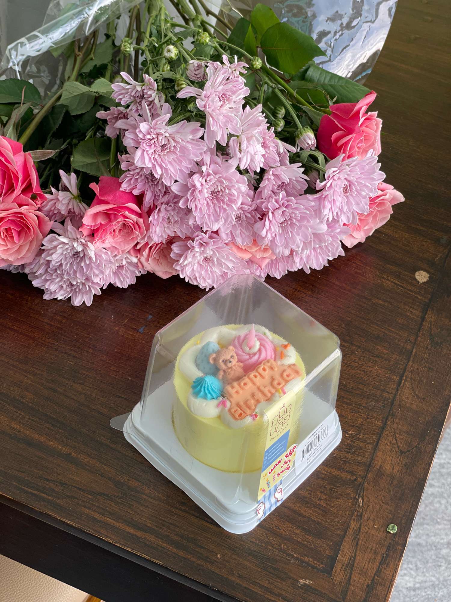 A bouquet of pink roses and chrysanthemums is arranged on a dark wooden table, with a small, pale yellow cake in a clear plastic container placed in the foreground. The cake is decorated with a teddy bear figurine, blue and pink frosting, and orange lettering that reads "Happy Birthday." A label on the cake container displays Thai script and graphics. The table surface shows natural wood grain and some minor imperfections. The background is blurred, suggesting an indoor setting, possibly in Nong Prue, Thailand, given the presence of Thai script on the cake packaging.