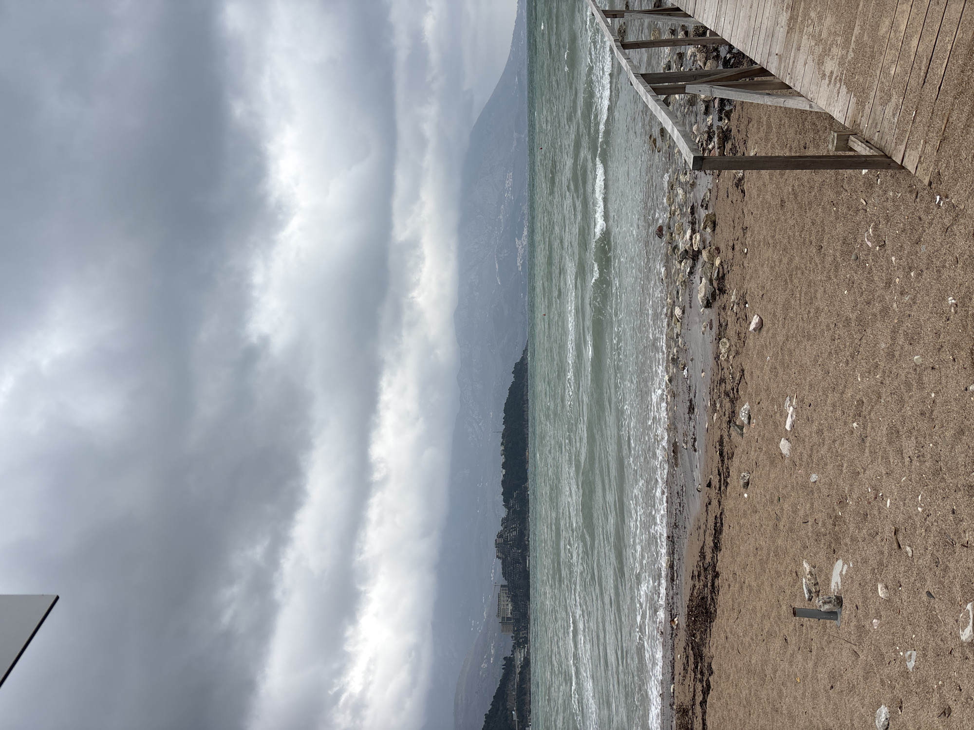 The image displays a turbulent coastal scene in Budva, Montenegro, viewed from a sandy beach looking east towards Bečići, with the distinctive Splendid Conference & Spa Resort complex discernible on a distant, vegetated headland. The sky is entirely covered by a uniform layer of dense grey clouds. The sea exhibits rough, dark green-blue waters with numerous white-capped waves breaking towards the shore. Two small red spherical buoys are visible on the water's surface in the mid-distance.
The foreground consists of a light brown sandy beach, scattered with small white and grey rocks, assorted debris, and a prominent accumulation of dark brown seaweed at the water's edge. A weathered wooden walkway or pier, constructed from planks and featuring a simple wooden handrail, extends diagonally from the lower right corner into the frame. In the left foreground, a grey metallic pipe or post protrudes vertically from the sand, surrounded by several larger, light-colored stones. The background features large mountains, their upper portions obscured by low-hanging clouds and atmospheric haze, with scattered light-colored structures visible on their lower slopes. No individuals are present in the scene.