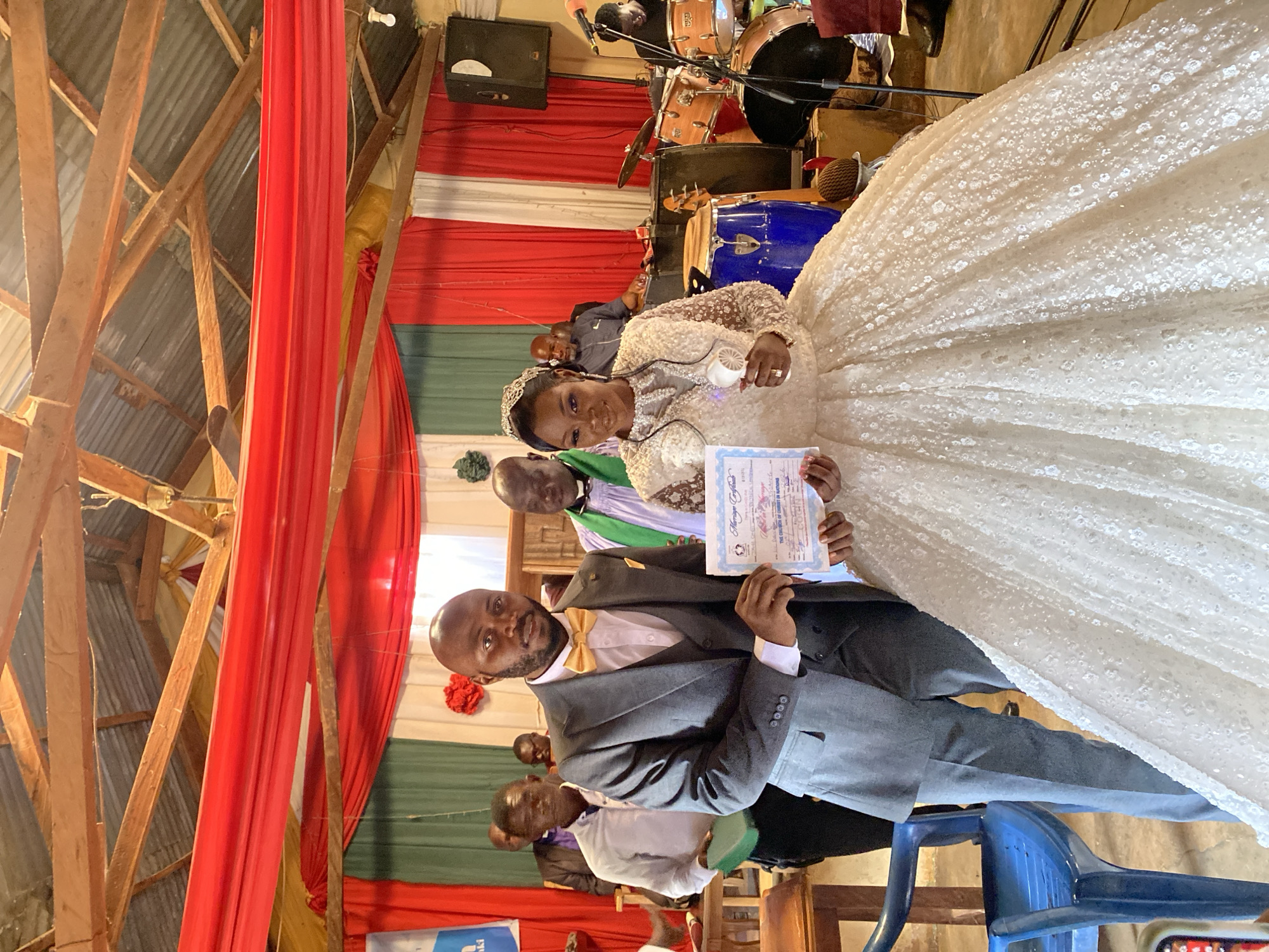 The image captures a wedding ceremony in Vom, Nigeria. A bride in a heavily embellished white gown with a tiara and a groom in a gray suit with a gold bow tie are holding a marriage certificate. They are surrounded by other people who appear to be wedding guests. The background features red, white, and green fabric decorations, along with a drum set and speaker setup. The interior has wooden beams, indicative of a simple building structure. A blue plastic chair is visible in the foreground.