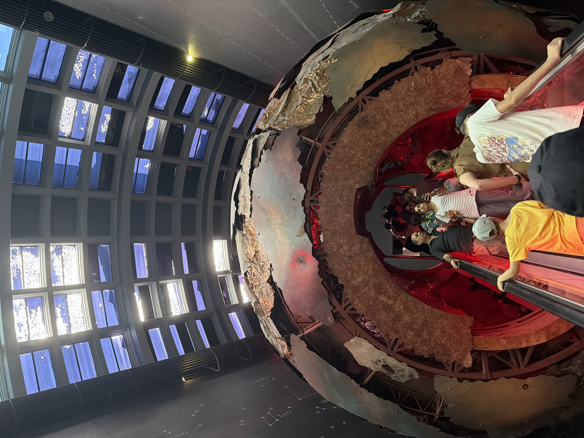 The image shows the inside of the Globe at the London Science Museum. There are people riding up and down an escalator inside the globe. The building's architecture includes multiple windows, which are visible in the background. There is a pattern of stars on the floor of the location.