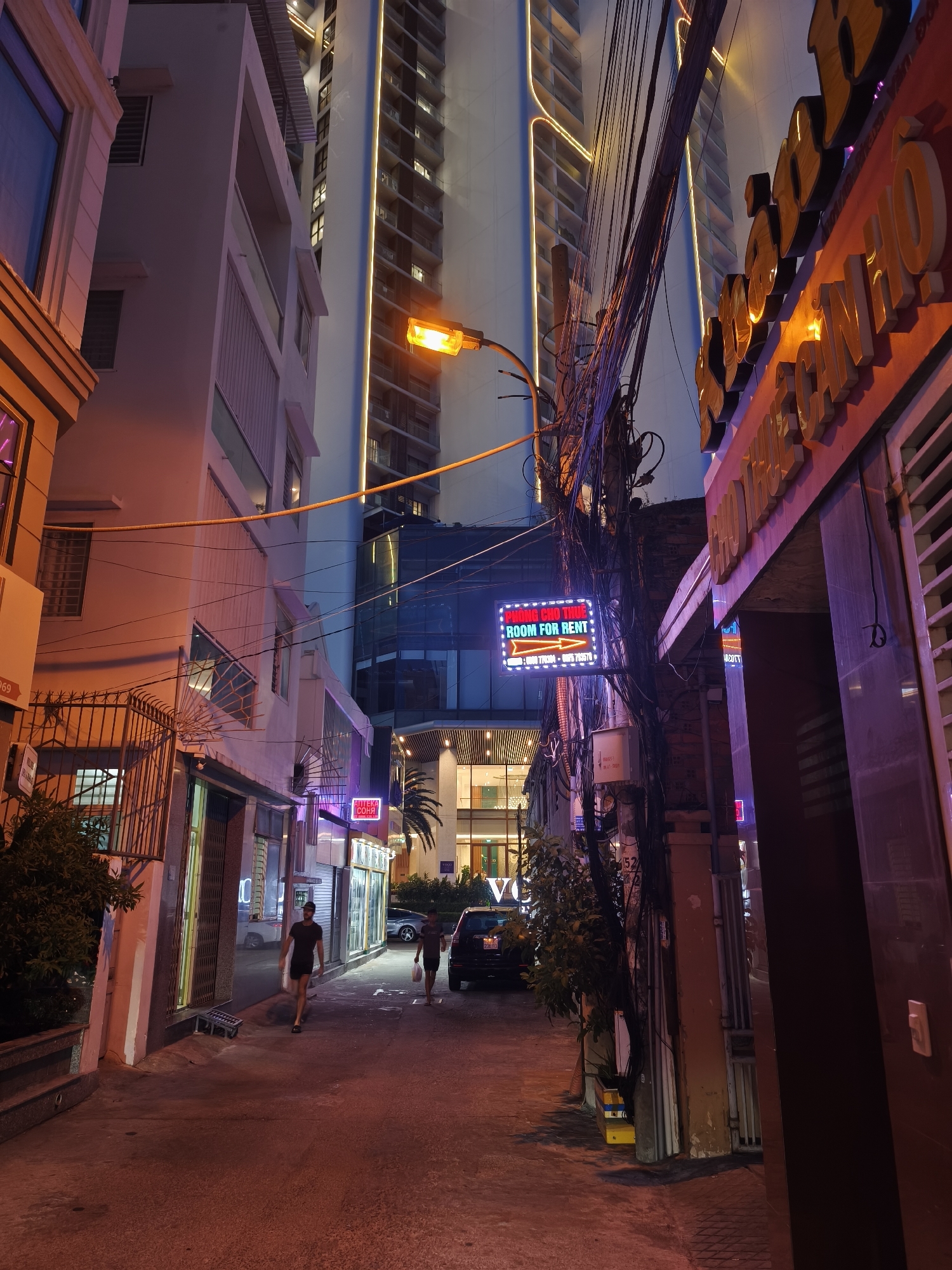 The image depicts a narrow alleyway in Bac Nha Trang Ward, Vietnam, at night. The scene is illuminated by streetlights and neon signs, creating a vibrant and somewhat chaotic atmosphere.
On the left side of the alley, a building with white facade and balconies rises, its windows lit from within. A metal fence with intricate designs borders the lower part of the building. Further down, a neon sign with Vietnamese characters is visible.
To the right, another building displays a large, illuminated sign with bold, three-dimensional lettering in a warm orange and yellow hue, also with Vietnamese text. A thick bundle of tangled electrical wires hangs overhead, adding to the urban feel.
In the middle of the alley, a streetlamp casts a bright orange glow. A neon sign with English text "ROOM FOR RENT" is mounted on a pole, featuring a small arrow pointing to the right.
Two men are walking in the alley. One, wearing a dark t-shirt and shorts, is walking towards the viewer. Another man, further down, is walking away from the viewer, carrying a plastic bag. A dark-colored car is parked on the right side of the alley.
The ground is paved with concrete, reflecting the colorful lights. The overall impression is of a lively, albeit somewhat cluttered, urban environment at night.