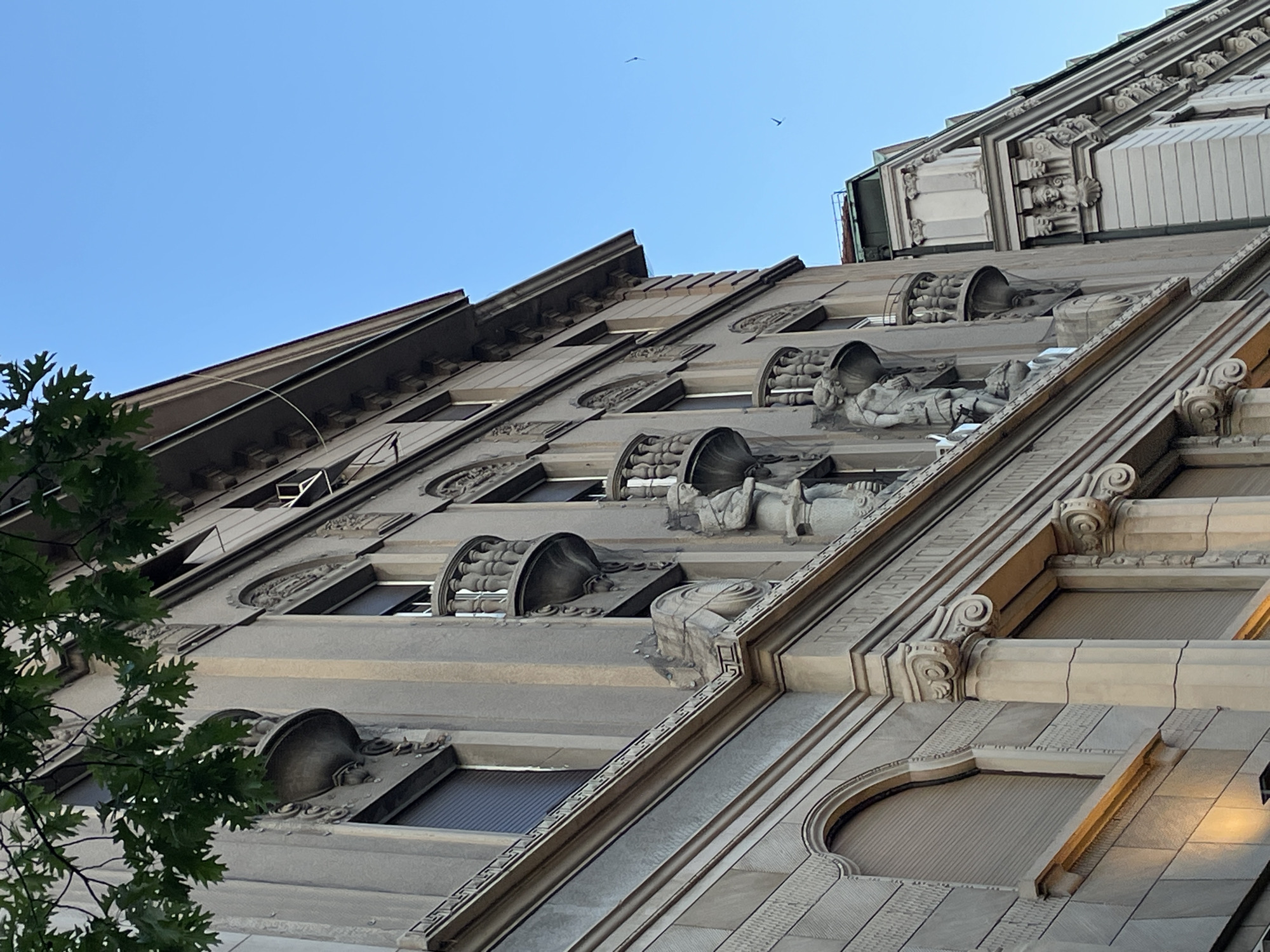 The image depicts a low-angle view of a building facade, exhibiting detailed architectural features such as window ornaments, sculpted figures, and intricate moldings. The building is constructed from a light-colored stone. The sky is clear and blue with two birds flying in the upper portion. A small part of a tree is visible in the lower left corner. The building is located somewhere outside of Belgrade, Serbia.