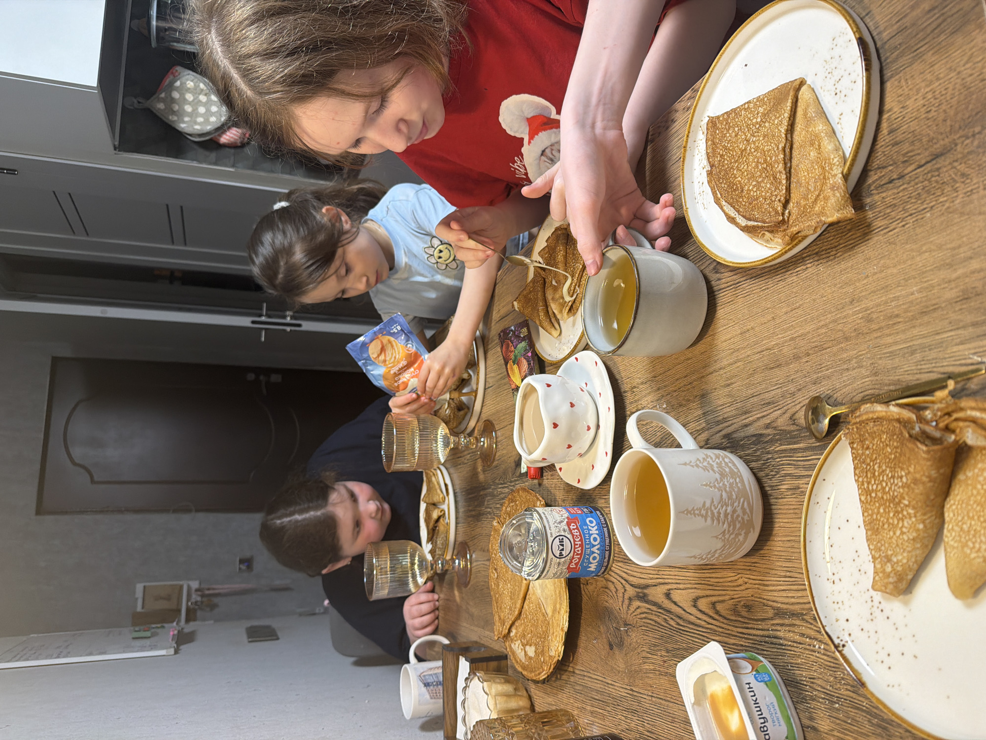 Three children sit at a wooden table laden with pancakes, condensed milk, and tea. A girl in a red shirt pours a golden sauce over a pancake, while a girl in a light blue shirt squeezes filling from a packet. A third child sits across the table with a glass of water. The table setting includes plates with pancakes, mugs of tea, and a can of condensed milk labeled in Russian. The location is likely a home kitchen. Given the labels on the food items, the city is Perm, Russia.