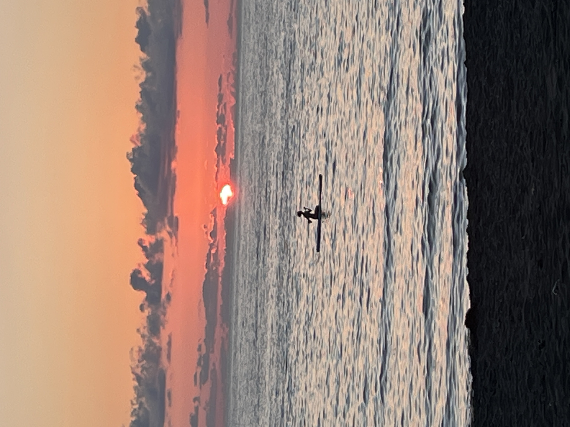 A solitary figure is paddleboarding on the Black Sea at sunset in Ureki, Georgia. The sun, a bright orb, is setting just above the horizon, casting a warm orange and pink glow across the sky and reflecting on the calm water. Clouds are scattered across the sky, tinged with the sunset colors. The foreground shows a dark, sandy beach. The paddleboarder is silhouetted against the vibrant sky, appearing small in the vast expanse of the sea.
