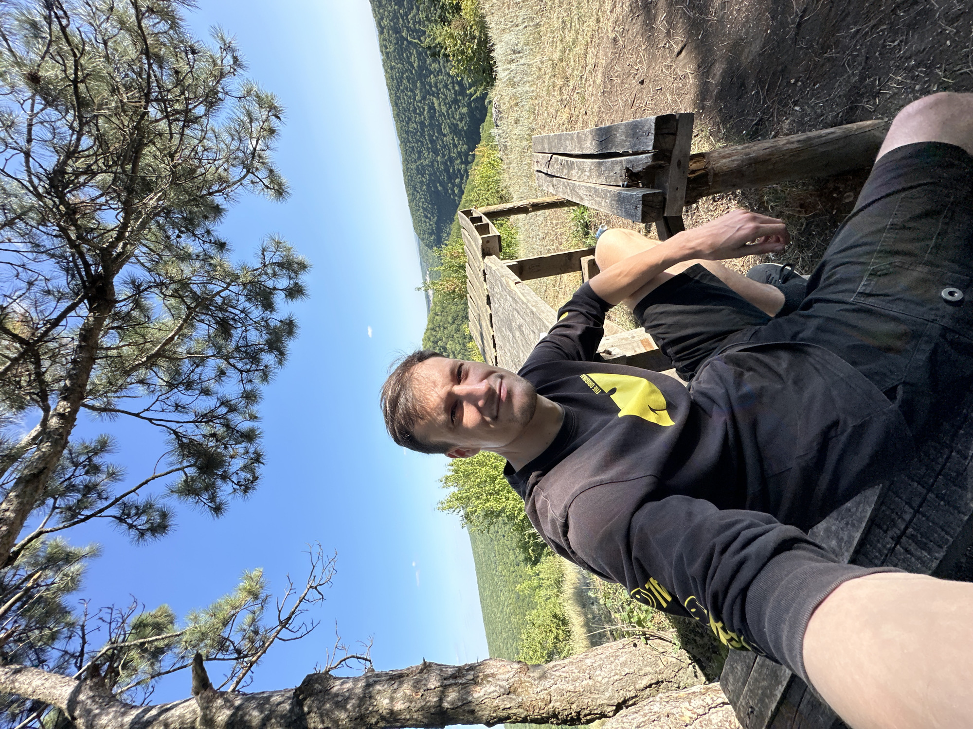 A man is taking a selfie outdoors, seated on a wooden bench next to a wooden table. He is wearing a black long-sleeved shirt with yellow smiley face graphics and black shorts. The man is looking towards the camera with a slight smile. Behind him, a lush green forest stretches across rolling hills under a clear blue sky. The scene appears to be in a rural or natural setting. The table and benches are weathered, suggesting they have been outdoors for some time. A large tree trunk is visible to the left of the man.