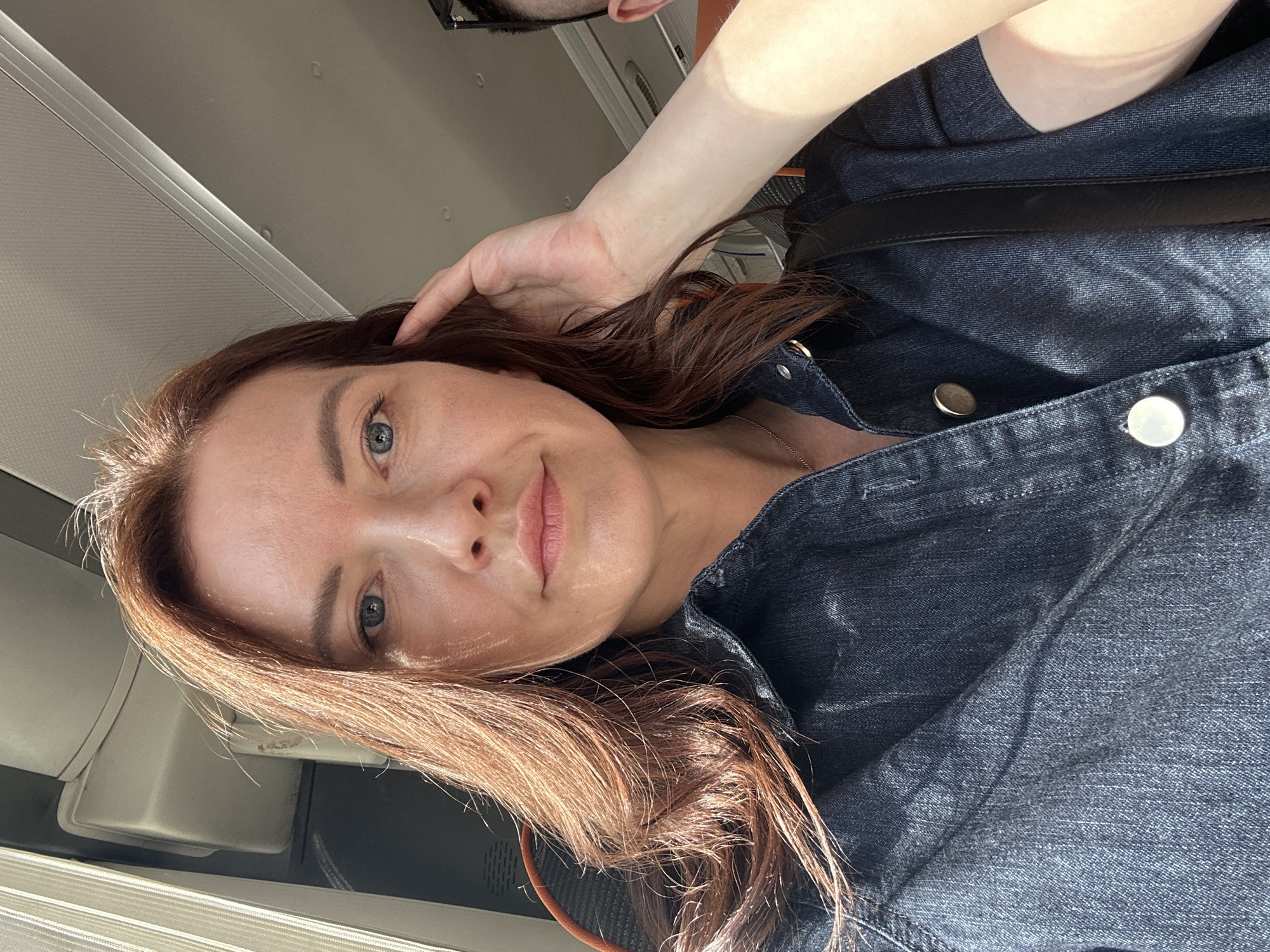 A selfie shows a fair-skinned woman with auburn hair and blue eyes, wearing a dark denim jacket with silver button closures. A black strap is visible over her right shoulder. Her left hand is touching her hair. The background appears to be an airplane interior with a window and overhead compartment.