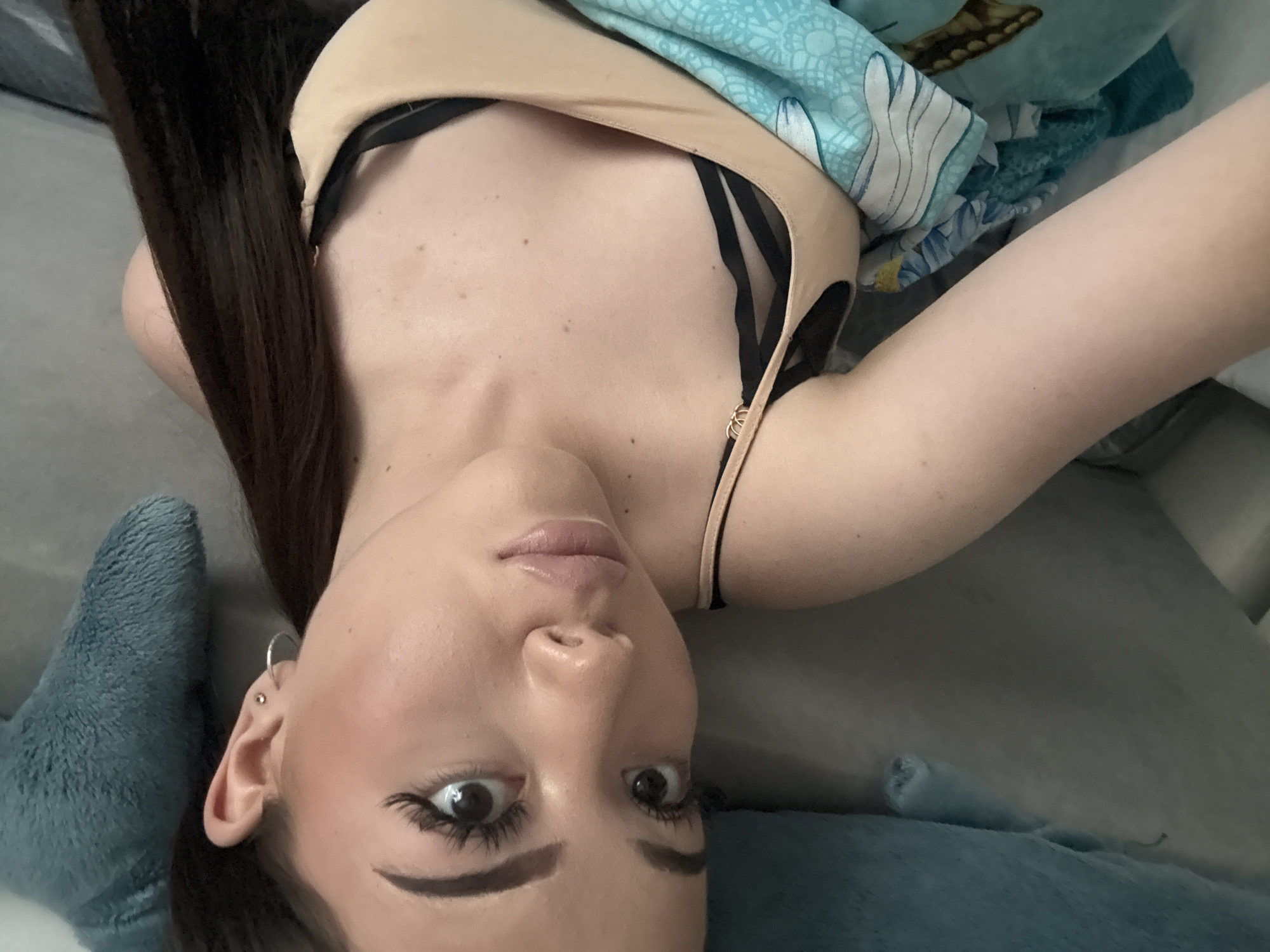 A woman with long brown hair, wearing a beige top with black straps, lies on a grey surface with a blue patterned blanket partially covering her. She has an earring in her left ear and is looking directly at the camera. A blue stuffed animal is positioned near her head. The location appears to be indoors. The image does not show any location information related to Naberezhnye Chelny, Russia.