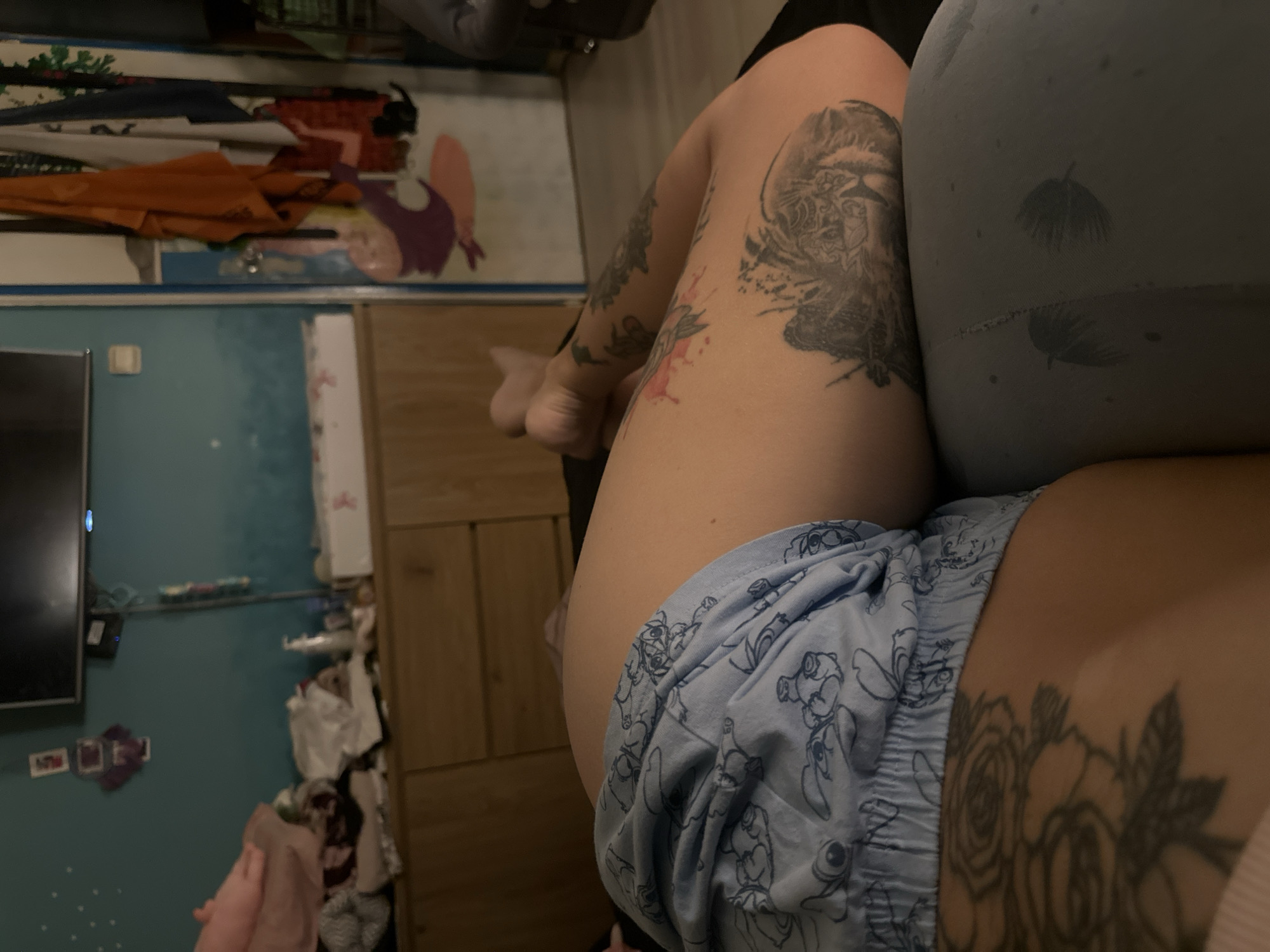 The image shows a close-up view of a person lying down, with their body partially visible. The person is wearing light blue shorts with a pattern of cartoon characters. They have several tattoos on their leg and arm, including a large tattoo of a tiger on their thigh and floral designs on their leg and arm. The background includes a dresser with various items on it, a television mounted on the wall, and a doorway with clothes hanging on it. The lighting is dim, suggesting the photo was taken indoors in the evening or at night. There are no discernible elements that would indicate a specific location within the city of Plovdiv, Bulgaria.