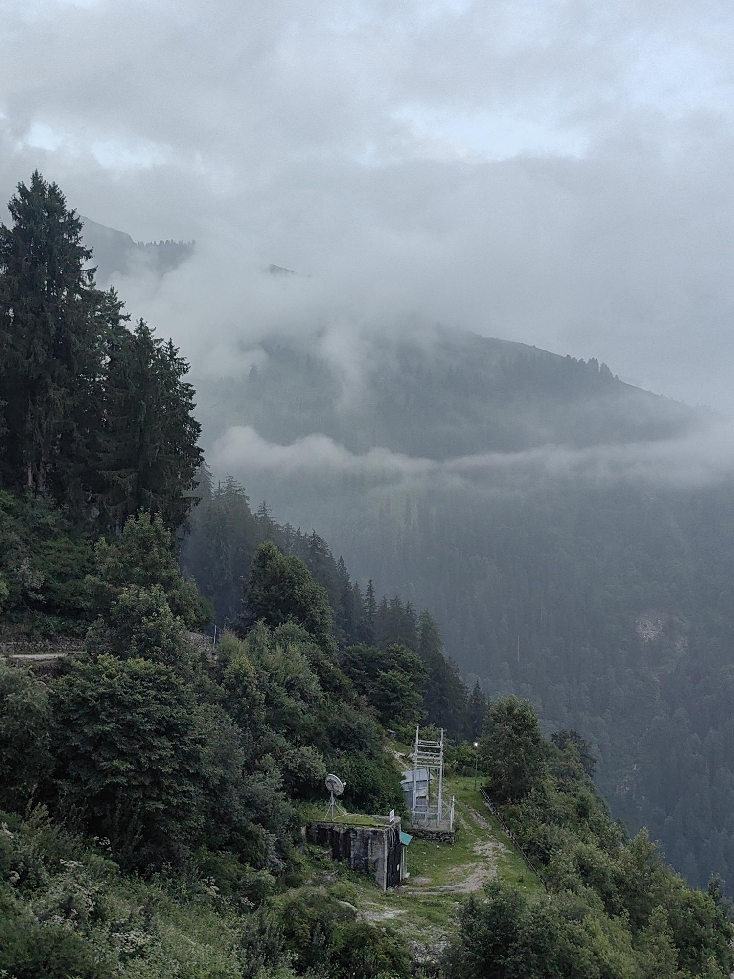 The image depicts a misty mountainous landscape with lush green vegetation. In the foreground, a steep, forested slope is visible, with a winding path leading upwards. A small, concrete structure with a metallic antenna stands on a raised platform amidst the greenery. Further up the slope, a more elaborate metal structure, possibly a tower or equipment housing, is situated near another building. Dense pine trees dominate the left side of the image, their dark foliage contrasting with the lighter greens of other trees and shrubs. The background is characterized by rolling hills covered in trees, partially obscured by thick fog and clouds, creating a serene and somewhat mysterious atmosphere. The scene suggests a remote, elevated location, likely within a mountainous region. This specific location is in Prini, India.