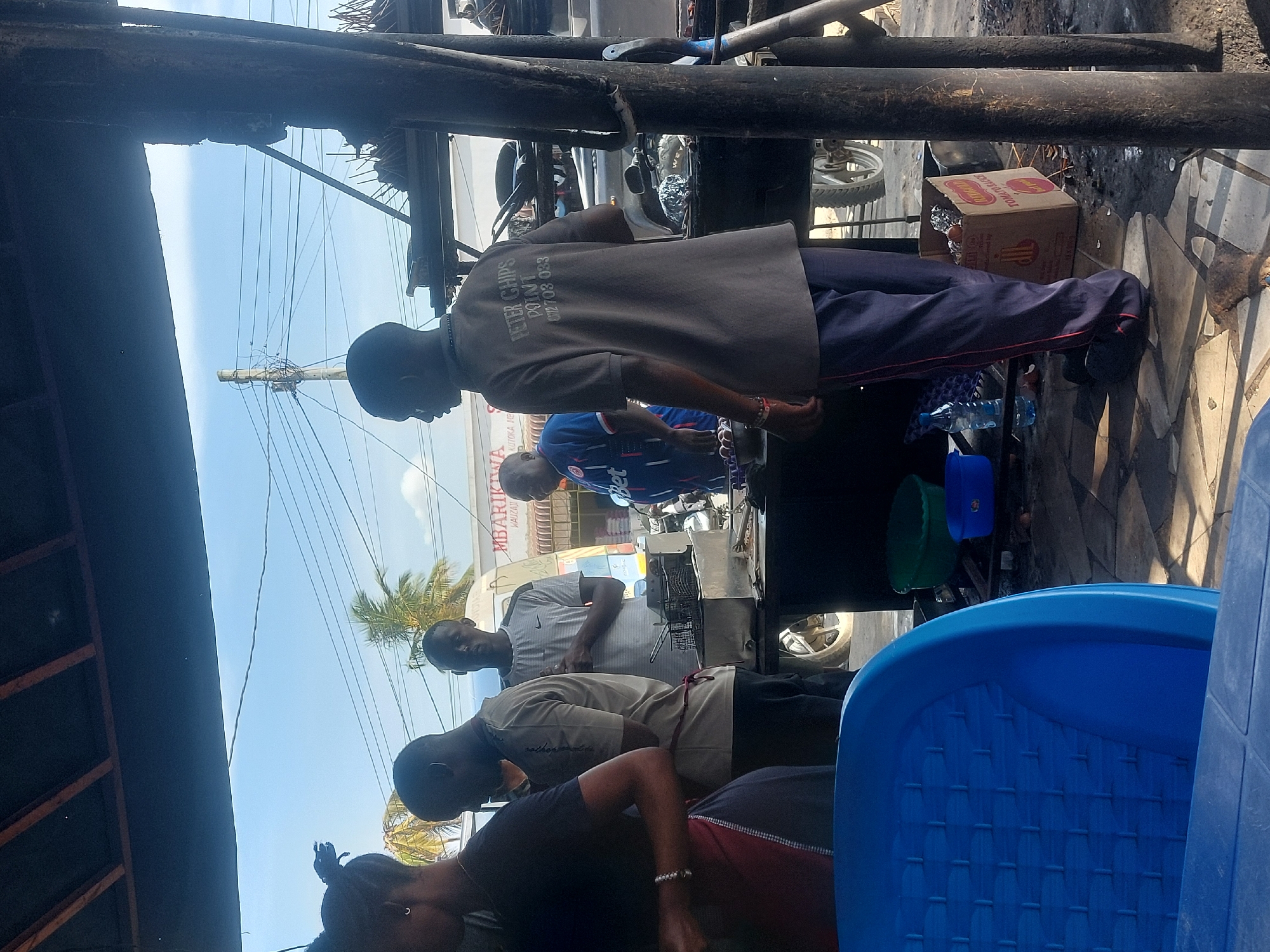 The image shows an outdoor food stall in Dar es-Salaam, Tanzania. Four people stand around a deep fryer, possibly preparing food. One person wears a shirt with "PETER CHIPS POINT 072703 033" printed on the back. A box, blue and green basins, and blue plastic chairs are nearby. In the background, a white van parked in front of a building with the partial word "MBARIK WA..." visible on its facade. Overhead power lines are visible against a clear sky with a palm tree in view. The scene appears to be an informal food vending setup.