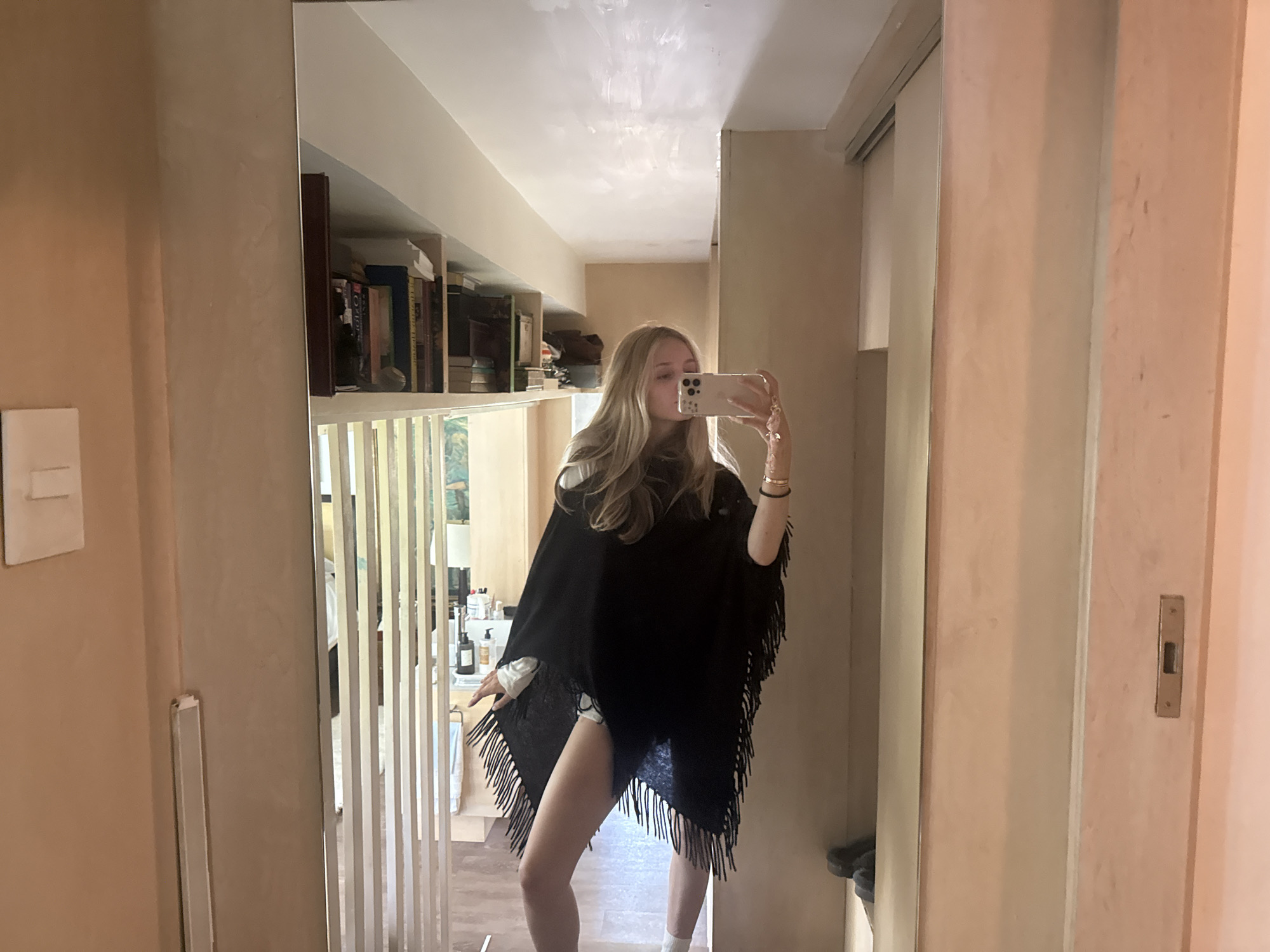 A young Caucasian woman with blonde hair is standing in a hallway, taking a selfie in a full-length mirror. She is wearing a black fringed poncho over a white shirt and white shorts. She is holding a gold-colored smartphone in her right hand. The hallway has light-colored walls and a wooden floor. To her left, there is a bookshelf filled with books. To her right, there is a sliding door. The scene appears to be inside a residential building in Mexico City, Mexico.