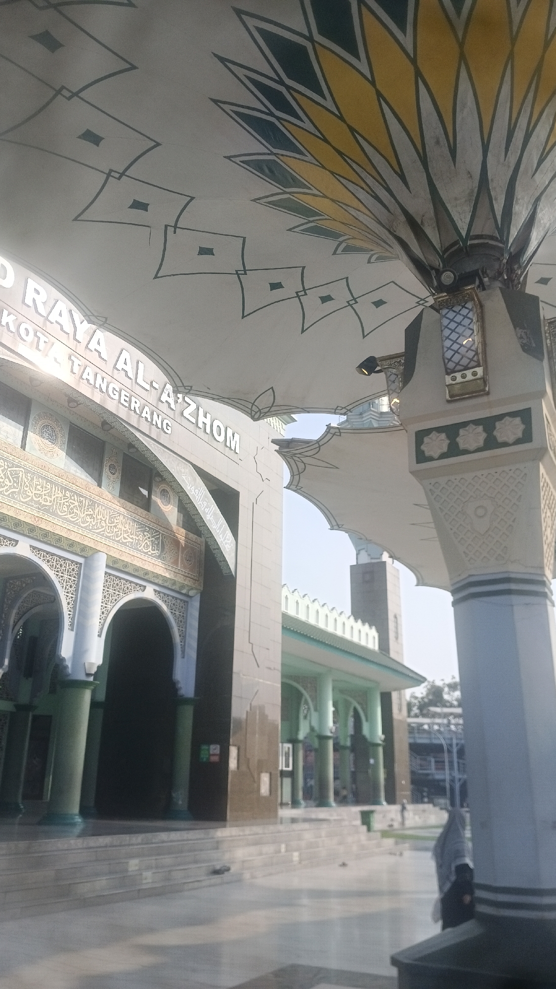 The image captures an exterior view of the Al-Azhom Mosque in Tangerang, Indonesia. The foreground features a large, white, tensile fabric structure with a decorative pattern of yellow and green geometric shapes, extending from a cylindrical column. To the left, the facade of the mosque is visible, with ornate Arabic calligraphy and traditional archways. The words "RAYA AL-AZHOM" and "KOTA TANGERANG" are prominently displayed on the building's exterior. In the background, another section of the mosque with green roofing and supporting pillars can be seen, along with a hint of trees and a distant walkway. A woman wearing a hijab is partially visible on the right side of the frame, walking away from the camera. The scene is illuminated by natural daylight, casting soft shadows.