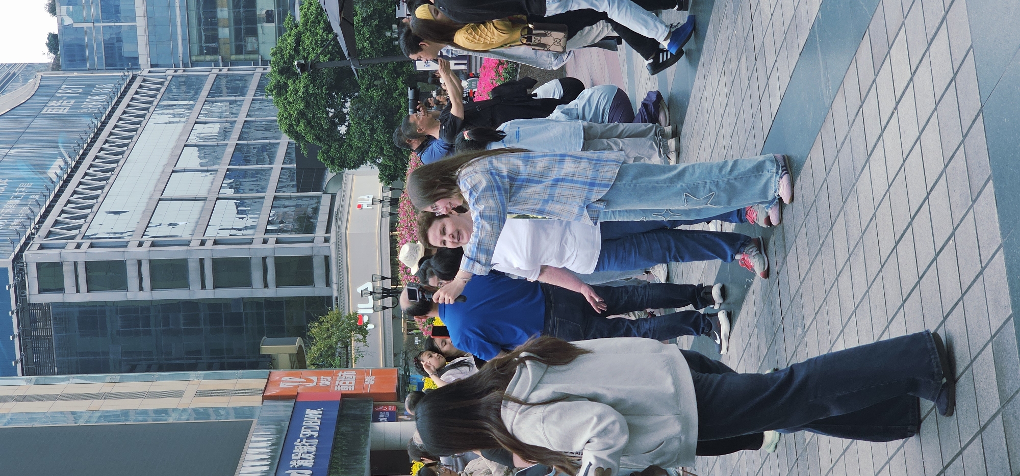 A photograph captures a scene in Yuzhong District, China, featuring a crowd of people primarily composed of Asian individuals. They are situated on a tiled pathway, some holding smartphones as if taking pictures or videos. A tall building with mirrored windows is visible in the background, while storefront signs written in Chinese appear along the lower part of the image. Many individuals are casually dressed. The general impression is a public space.