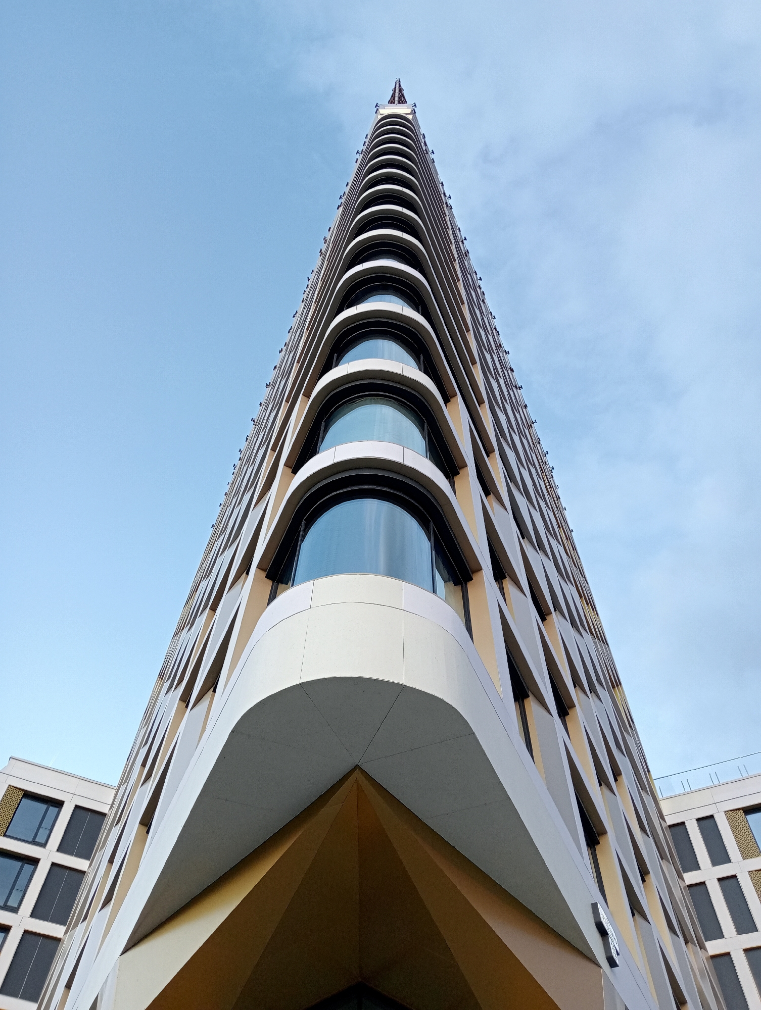 The image shows a low-angle shot of the modern, angular facade of a tall building against a cloudy sky. The building's exterior features a repeating pattern of diagonal lines and recessed windows with curved tops. The corner of the building is sharply pointed, creating a dramatic perspective. To the left, another building with a grid-like window pattern is partially visible. The building appears to be the Bonalumi residential complex, located in Saint Petersburg, Russia.