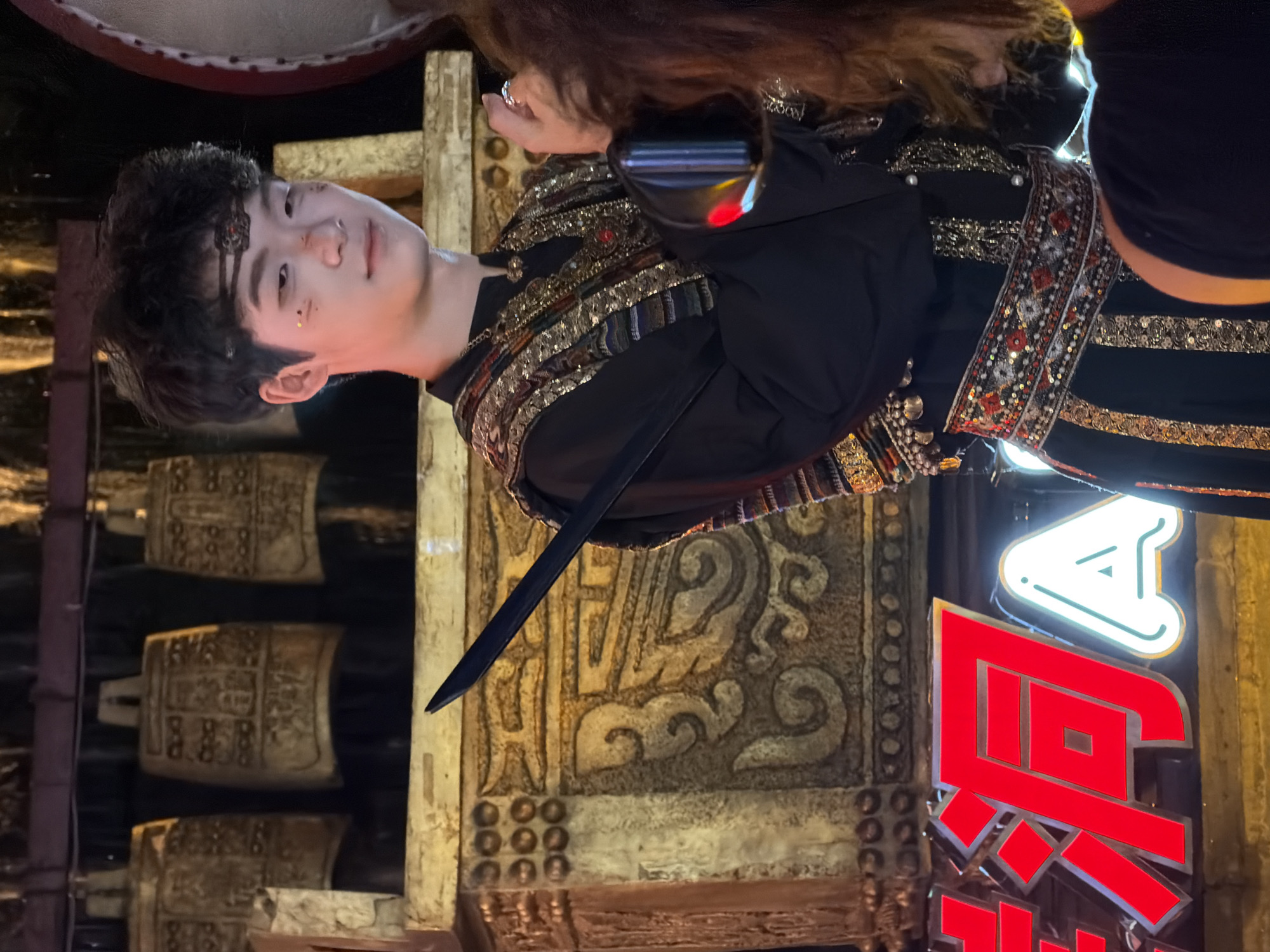 In Yuzhong District, China, a young man with dark, curly hair and a headband adorned with red and black beads is depicted. He has light skin and a smattering of gold glitter on his cheeks and nose. He is wearing a black garment with intricate gold and multi-colored embroidery across the chest and a bejeweled belt. A dark, thin sword is held diagonally across his torso. Behind him, a row of decorative, ancient-looking bronze bells hangs, and elaborate golden ornamentation adorns a structure. In the foreground, a portion of a large, bright red Chinese character and a white illuminated letter "A" are visible, suggesting a neon sign or advertisement. The overall scene appears to be set at night, possibly during a cultural performance or festival.
