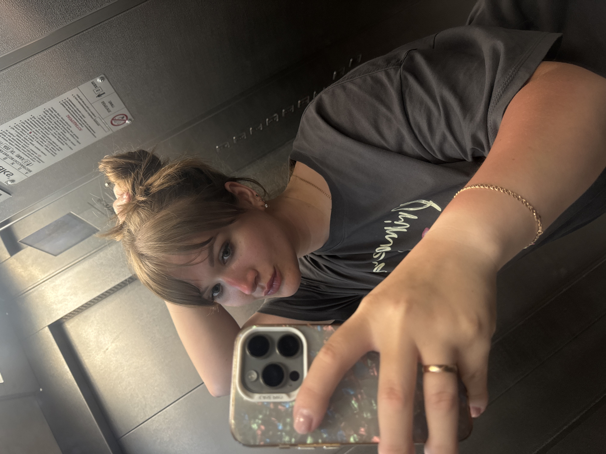 A person with light brown hair pulled up in a messy bun is taking a selfie in an elevator. The person is wearing a dark gray T-shirt and gold jewelry, including a necklace and bracelet. The phone case is patterned with a marbled design. The elevator walls are gray metal, with a sign on one wall displaying text in a Cyrillic alphabet. The image appears to have been taken inside an elevator in Yevpatoriya, Ukraine.