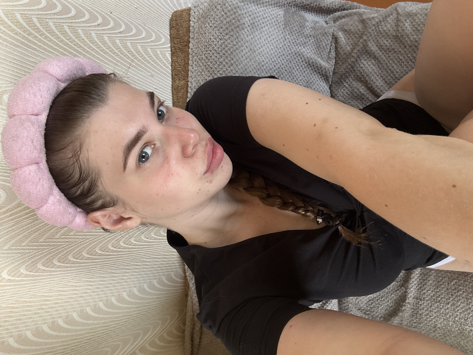A young woman with a pink headband is lying on a patterned beige couch. She has light skin, blue eyes, and her hair is braided. She is wearing a black shirt. The background includes a textured gray blanket. There is no information to indicate this photograph was taken in or relates to the city of Meshchery, Russia.