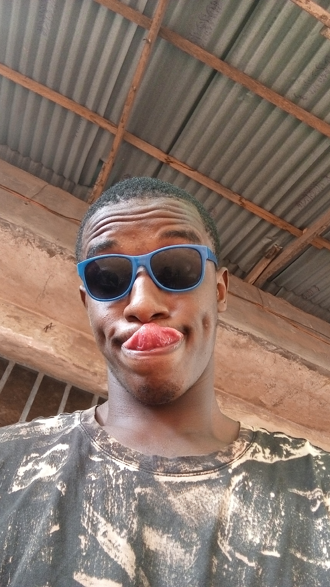 A young person is captured in a close-up, slightly low-angle portrait, looking directly at the camera with a playful expression. They have dark skin, short dark hair, and prominent dimples on both cheeks. Their lips are pursed, with the tongue visible, adding to the quirky and lighthearted demeanor. They are wearing bright blue wayfarer-style sunglasses, which cover their eyes.
The individual is wearing a dark t-shirt with a faded, bleached, or tie-dye pattern in lighter shades, primarily beige or off-white, creating an abstract design.
The background primarily features the underside of a corrugated metal roof, common in many parts of Nigeria. Several wooden beams run horizontally and diagonally across the roof structure, providing support. To the left and slightly behind the person, a concrete or earth-colored wall is visible. The lighting suggests it is daytime, casting a soft, even illumination on the subject. There is no visible text in the image. The overall scene suggests a casual setting, possibly outdoors or in a sheltered open-air area consistent with the context of Onitsha, Nigeria.
