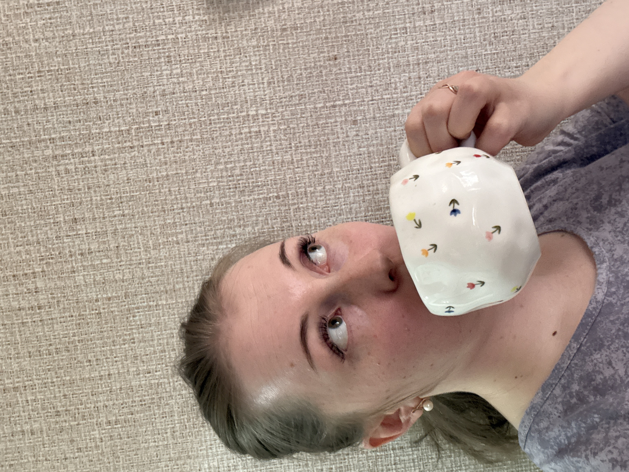 A close-up, slightly overhead shot shows a person, likely female, lying down on a textured, light beige surface, possibly a carpet or sofa. The person is oriented horizontally in the frame, with their head to the left and body extending to the right. They are holding a white ceramic mug with colorful, minimalist floral or arrow designs near their mouth.
The person is wearing a gray top with a subtle pattern and a small, delicate ring on their right hand's ring finger. A pearl earring is visible on their right earlobe. Their eyes are looking upwards, suggesting a moment of thought or looking at something above them, while their mouth is slightly open. Long, dark eyelashes are prominent. Their hair appears dark and is pulled back, partially visible around their head. The lighting suggests an indoor setting, likely natural light but not strong direct sunlight. There is no visible text in the image.