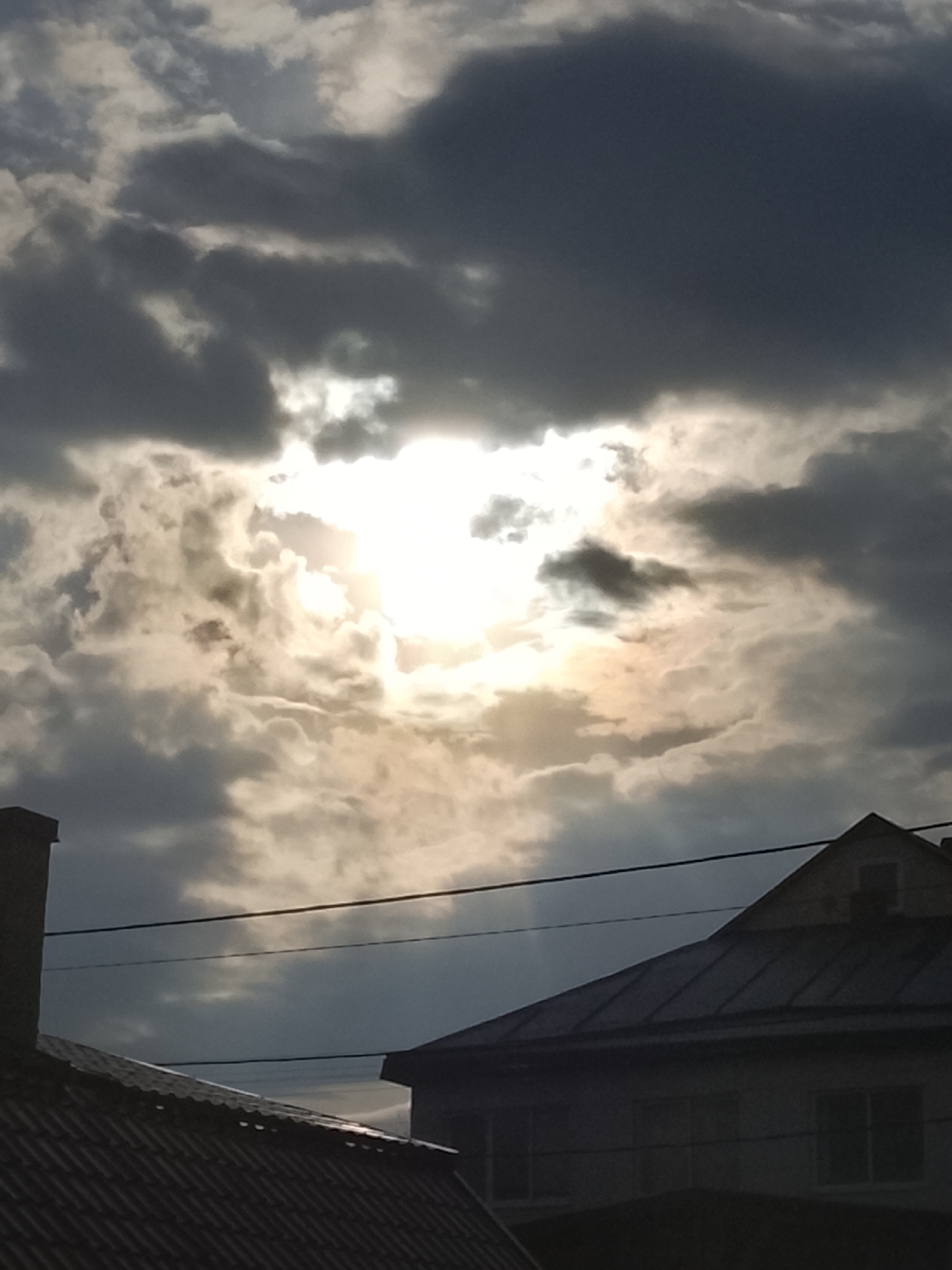 The image depicts a cloudy sky with the sun shining through the clouds. Below the sky, the roofs of two buildings are visible. The buildings are located in Chaikovsky, Russia. One building appears to have windows and an attic. Wires run horizontally across the frame.