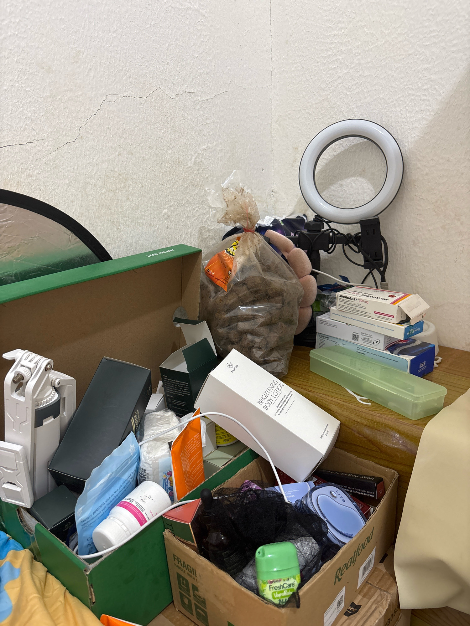 The image depicts a cluttered scene inside a room, likely a bedroom, in Wanajaya, Indonesia. The room is characterized by a white, textured wall with visible cracks. A ring light is mounted on a tripod, positioned on a wooden surface alongside various personal items and medication boxes. The medication includes Microgest 200 mg and other boxed pharmaceuticals.
Among the clutter, there are several cardboard boxes, some open and spilling their contents. Visible items include a white body lotion bottle, plastic packaging, a green plastic container, and a large bag filled with what appears to be dried food items or snacks. A white camera mount or accessory is also present. The floor is partially visible, showing bedding with a yellow and blue striped pattern. The overall impression is one of disarray and a lack of organization. There is no indication of any people or actions taking place in the scene.