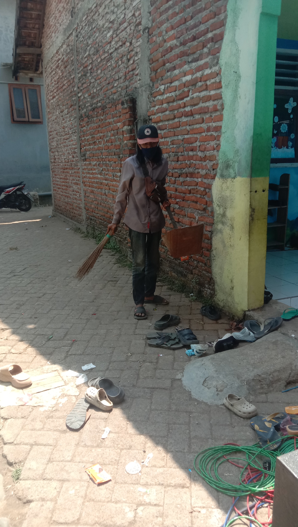A person, wearing a face mask, a baseball cap, a light brown button-down shirt, and dark pants, stands in an alleyway. They are holding a broom in their left hand and a dustpan in their right hand, appearing to be sweeping. Several pairs of sandals and flip-flops are scattered on the paved ground in front of them, some near a concrete structure on the right side of the image. A motorcycle is parked on the left side of the alley, against a brick wall. The alleyway is lined with brick walls on both sides, with a section of wall on the right painted with green and yellow accents and featuring a colorful mural or decoration. The ground is paved with rectangular stones. The scene is lit by sunlight, casting shadows. The location is identified as Karang Anyar, Indonesia.