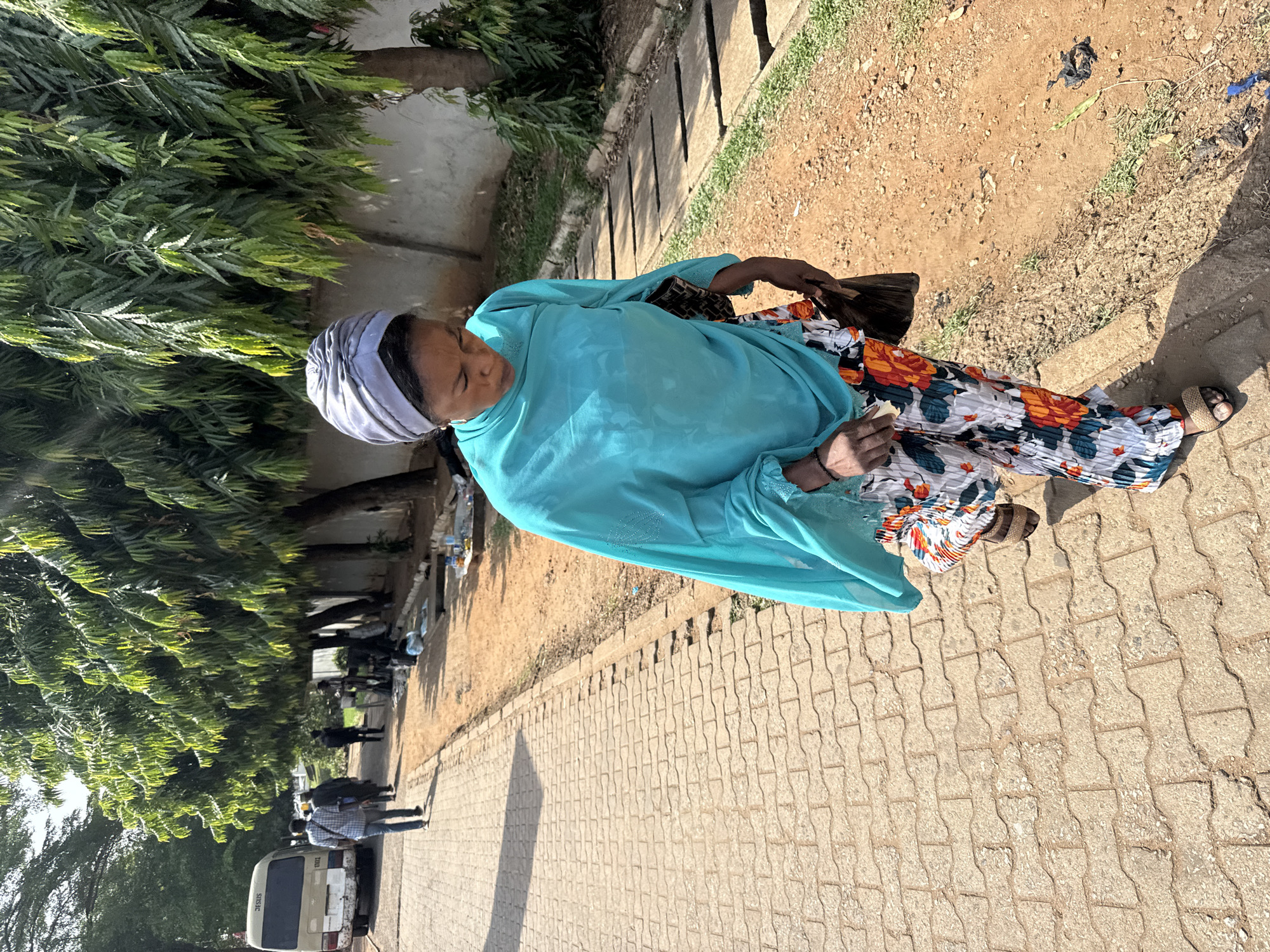 An elderly woman in Abuja, Nigeria stands on a brick-paved sidewalk next to a tree-lined road. She is dressed in a turquoise shawl and floral-patterned pants, wearing a gray head wrap and sandals. She carries a dark bag and holds something in her hand. Other people and a white van are visible further down the road. The surrounding ground is a mix of brick, dirt, and grass.