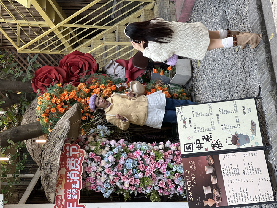 In Liangjiang, China, an outdoor scene depicts two East Asian females engaged in photography in front of a decorated establishment. The woman on the left, posing, wears a light yellow jacket, a white top or dress, dark blue pants, and a purple beret. She carries a small, light-colored bucket bag on her left shoulder and holds its strap with her right hand, looking towards her left. She stands before a backdrop consisting of dense artificial pink, light blue, and white roses, topped by a red sign with Chinese characters and a straw canopy. To her right, additional artificial orange roses and two large red artificial roses are displayed. A tree trunk rises between the floral arrangements. The woman on the right, viewed from behind, is holding a black smartphone horizontally with both hands, aiming it at the posing woman to take a photograph. She has long dark hair and wears a white cable-knit sweater dress, white knee-high socks, and light brown wedge boots, carrying a small purple quilted shoulder bag. To the far right, a bright yellow metal staircase ascends. The ground is covered with light grey gravel. In the foreground, two A-frame signs display menus in Chinese, one titled "Basque Series" with English text and images of desserts, the other titled "围炉煮茶" (Stove Tea) featuring an illustration of a teapot.