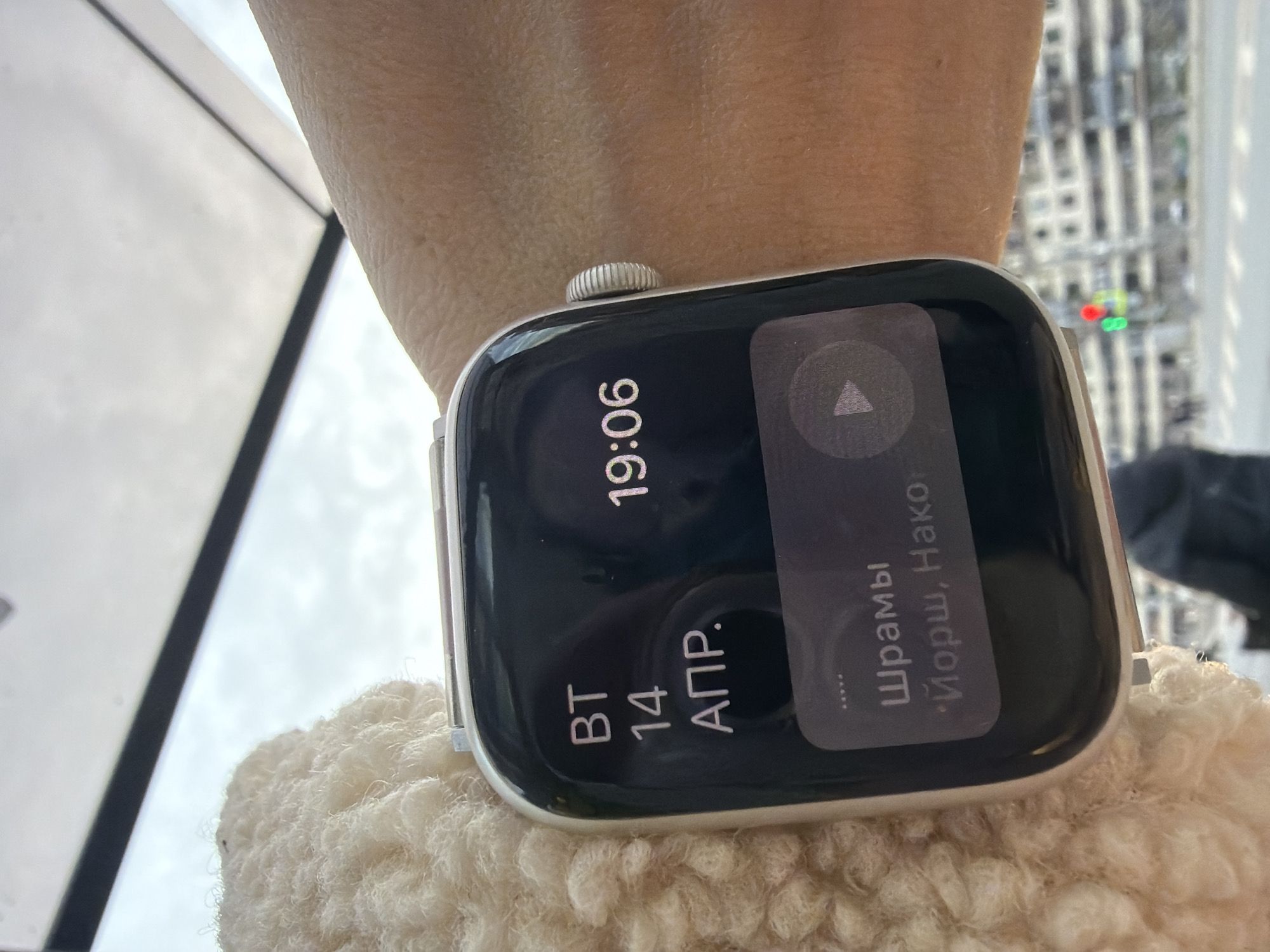 A close-up shot shows an Apple Watch on a person's wrist. The watch face displays the time "19:06" and the date "BT 14 Apr." A notification is visible on the watch, displaying a play symbol and the text "Шрамы • Йорш, Нако."
The background suggests a cityscape through a window, partially obscured and out of focus. Given that the text on the watch face is in Russian, the location can be inferred to be in Russia. Without specific landmarks visible in the out-of-focus background, pinpointing the exact location in Saint Petersburg is not possible.