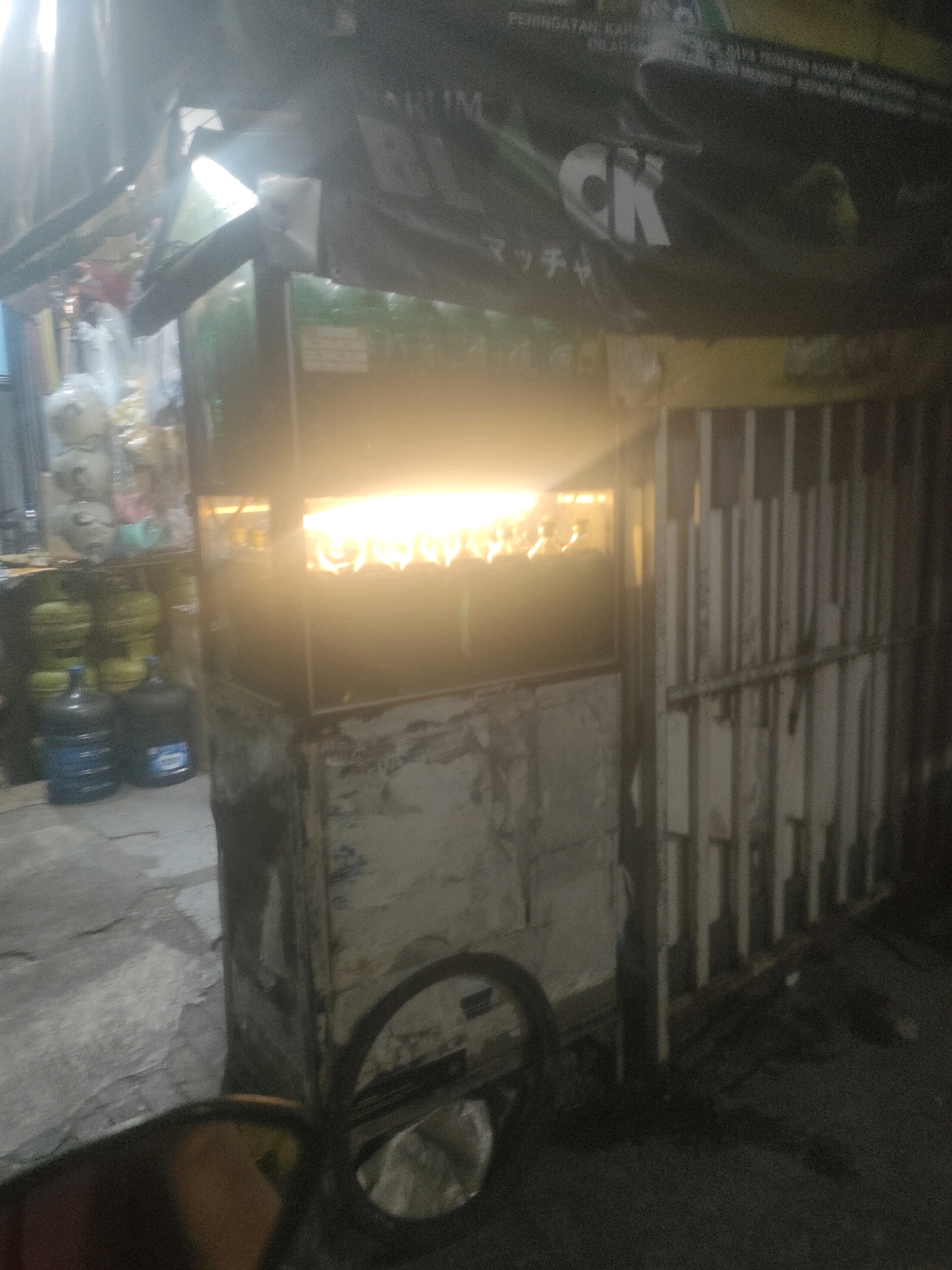 The image captures a food cart at night, likely in Surabaya, Indonesia, based on the Indonesian text visible on a banner. The cart is illuminated by bright lights from within, suggesting food is being prepared or displayed. A wheel is visible in the foreground, indicating the cart is mobile. In the background, several large water jugs and gas cylinders are stacked, commonly found in Indonesian street food setups. The overall scene is dimly lit, characteristic of a street scene at night. The banner on the cart has some text that appears to be a brand name or product advertisement. The lighting in the image is harsh, creating reflections and overexposure in some areas, which makes it difficult to discern fine details. The environment appears to be an outdoor urban setting.