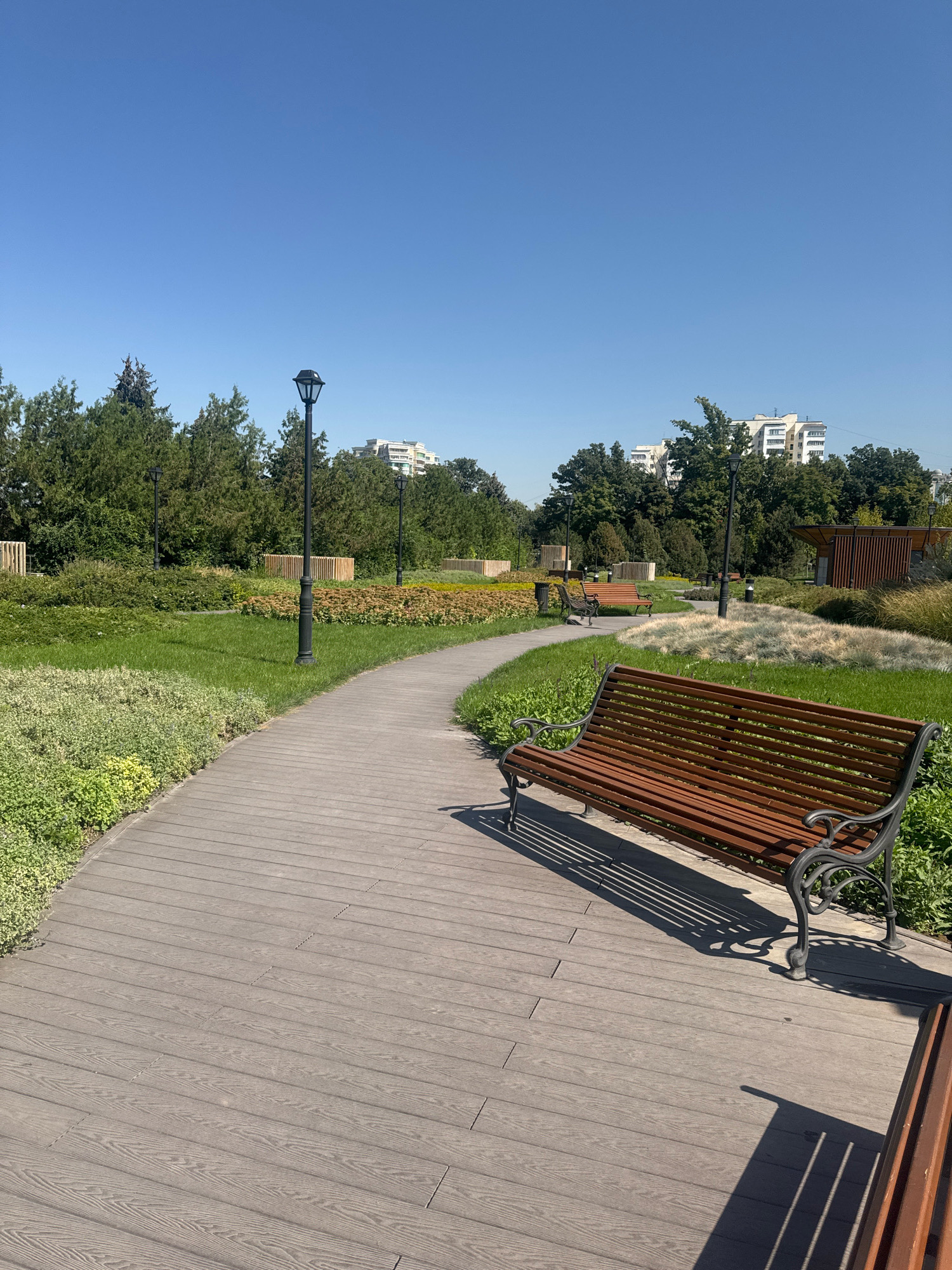 The image depicts a park scene on a clear, sunny day. A wooden pathway curves through the park, bordered by manicured green grass and various types of shrubs and plants. Several park benches are visible, one prominently in the foreground, with decorative metal legs and wooden slats. Lampposts are strategically placed along the pathway, and in the background, there are trees and modern residential buildings. The park appears well-maintained and offers a tranquil setting. The architecture of the buildings in the background suggests an urban environment, possibly in Almaty, Kazakhstan, given the context provided. There are no people in the image.