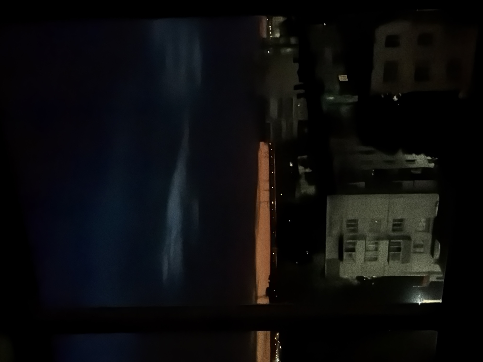 A nighttime view from a high vantage point shows a body of water, possibly the Kama River, on the left. A lit-up embankment runs along the water's edge. On the right, there is a section of the city of Chaikovsky, Russia, featuring several buildings with illuminated windows. The image appears to be taken from inside a building, looking out through a window frame.