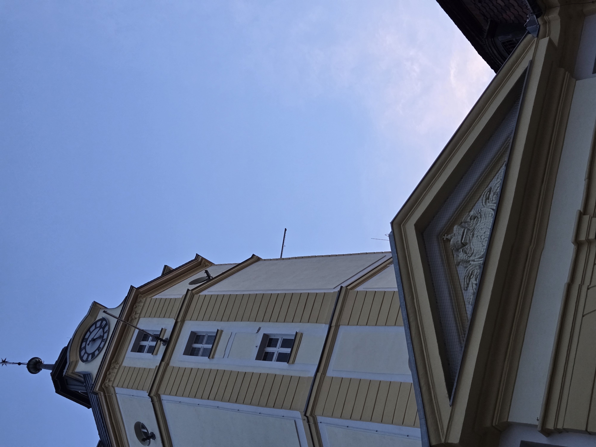 The image shows a close-up, low-angle view of a clock tower in Ansbach, Germany, against a clear blue sky. The tower, painted in cream and yellow, features several windows and architectural details. At the top of the tower, a clock face with Roman numerals is visible. A flagpole extends from the side of the tower. Below the tower, a section of a building with a triangular pediment and a carved coat of arms is visible. The overall scene is that of an historic building in an urban setting.