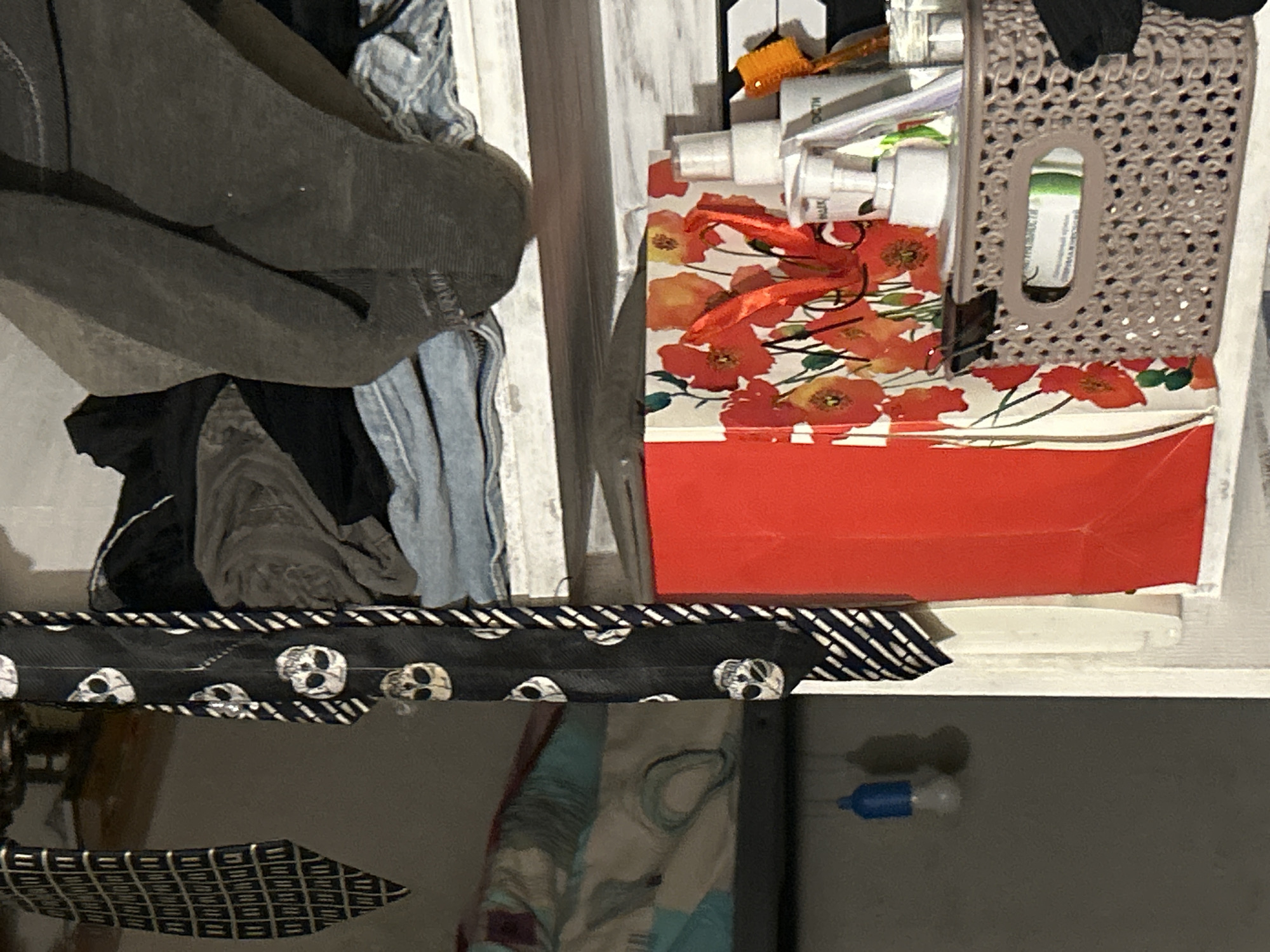 The image shows a cluttered shelf or storage area. There is a gray grater-like object, a red and white gift bag with floral patterns, and a black tie with a skull pattern. In the background, there are clothes or fabrics in shades of gray and blue. A tie with a geometric pattern is also visible. The environment appears to be indoors. The location is not discernible.