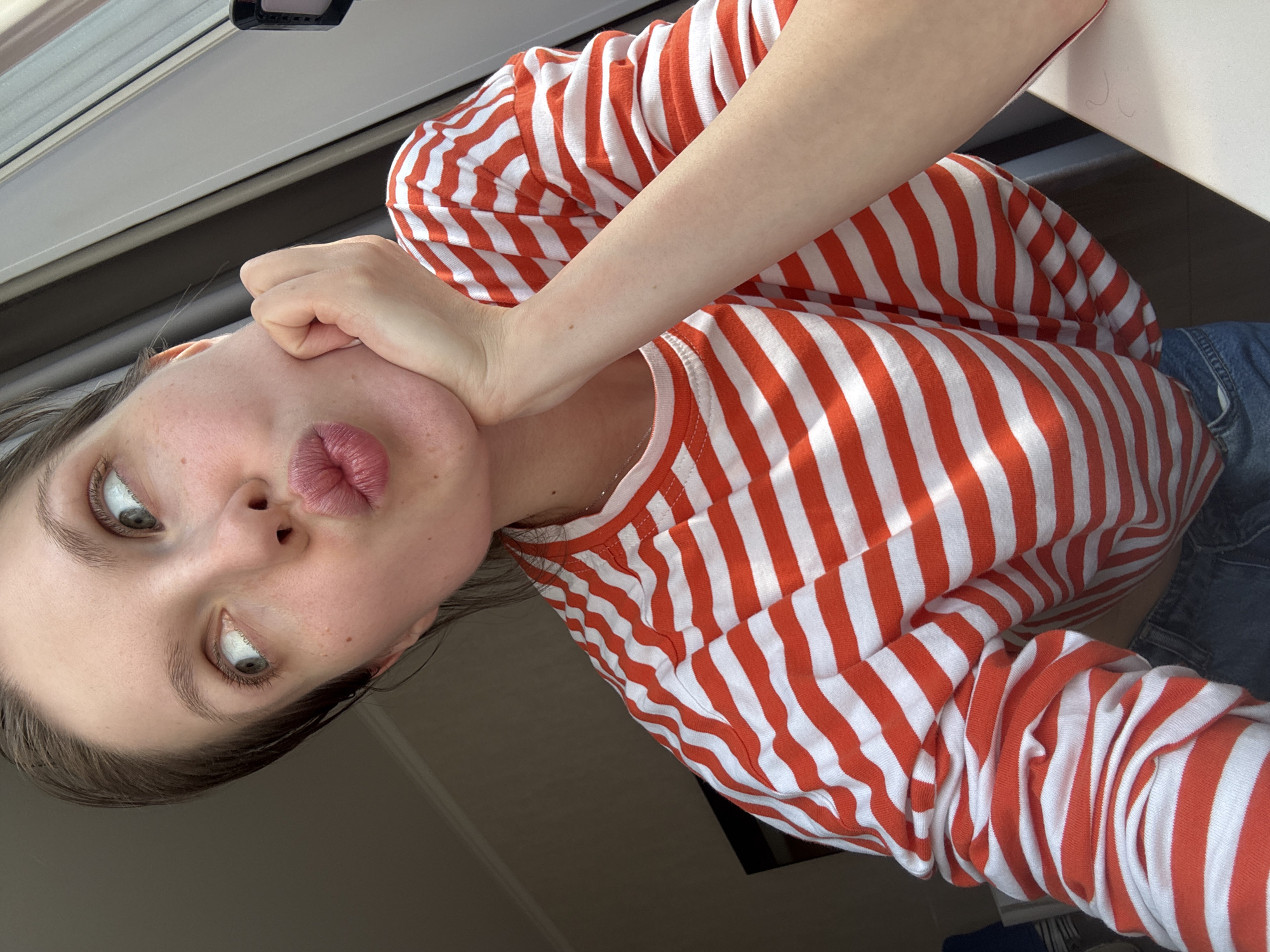 A young woman with light skin and grey eyes is visible in a close-up, slightly tilted selfie. She's wearing a long-sleeved shirt with horizontal alternating orange and white stripes. Her arm is positioned against her face, with her hand touching her cheek. Her lips are pursed into a pout. The background is partially obscured, revealing a window frame and neutral-toned walls. The image does not provide enough information to determine the location, let alone specify Moscow, Russia.