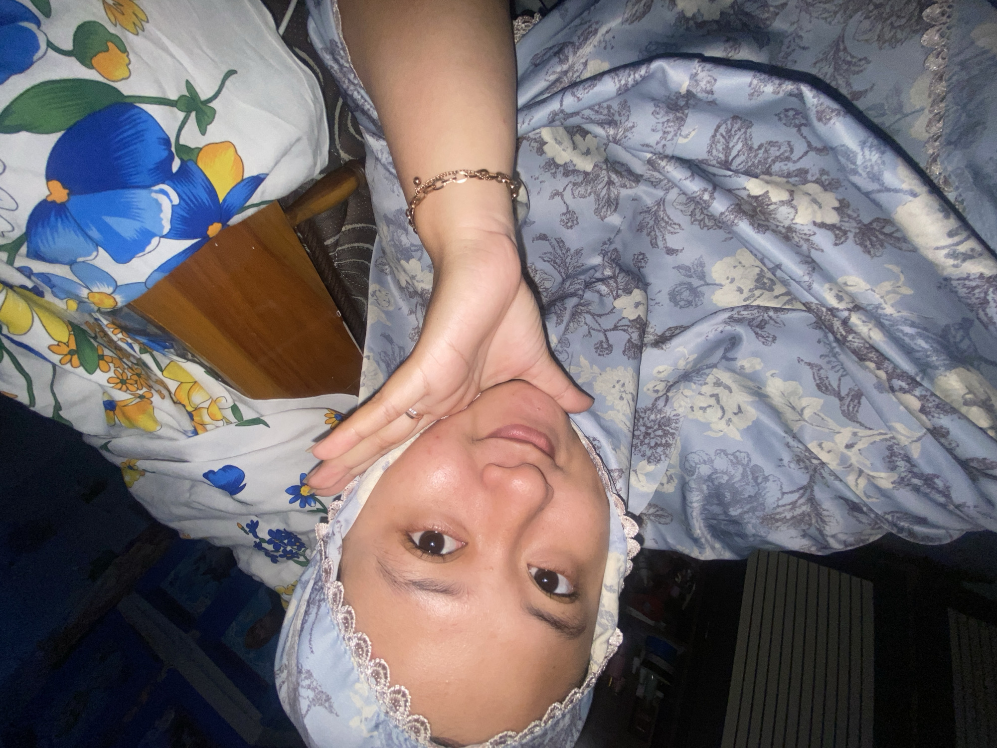 A young Indonesian woman, appearing to be in her late teens or early twenties, is pictured from a high-angle selfie perspective. She is wearing a light blue hijab with delicate white lace trim and a matching blue garment adorned with a floral pattern of white and dark blue flowers. Her right hand is raised, with her thumb and index finger touching her cheek and temple, creating a gesture that could be interpreted as playful or contemplative. A delicate, rose gold-colored bracelet with chain links and small charms is visible on her wrist. Her skin is smooth and glowing, and she has dark brown eyes and full lips. She is smiling slightly, looking directly at the camera. The background is dimly lit, with some patterned fabric visible, possibly bedding or curtains, featuring a white base with large blue and yellow flowers. There's also a glimpse of a wooden object, potentially a table or furniture. A dark, textured surface, possibly a mattress or carpet, is visible beneath the fabric. The scene appears to be indoors, likely in a bedroom. The image was taken in Gelam, Indonesia.
