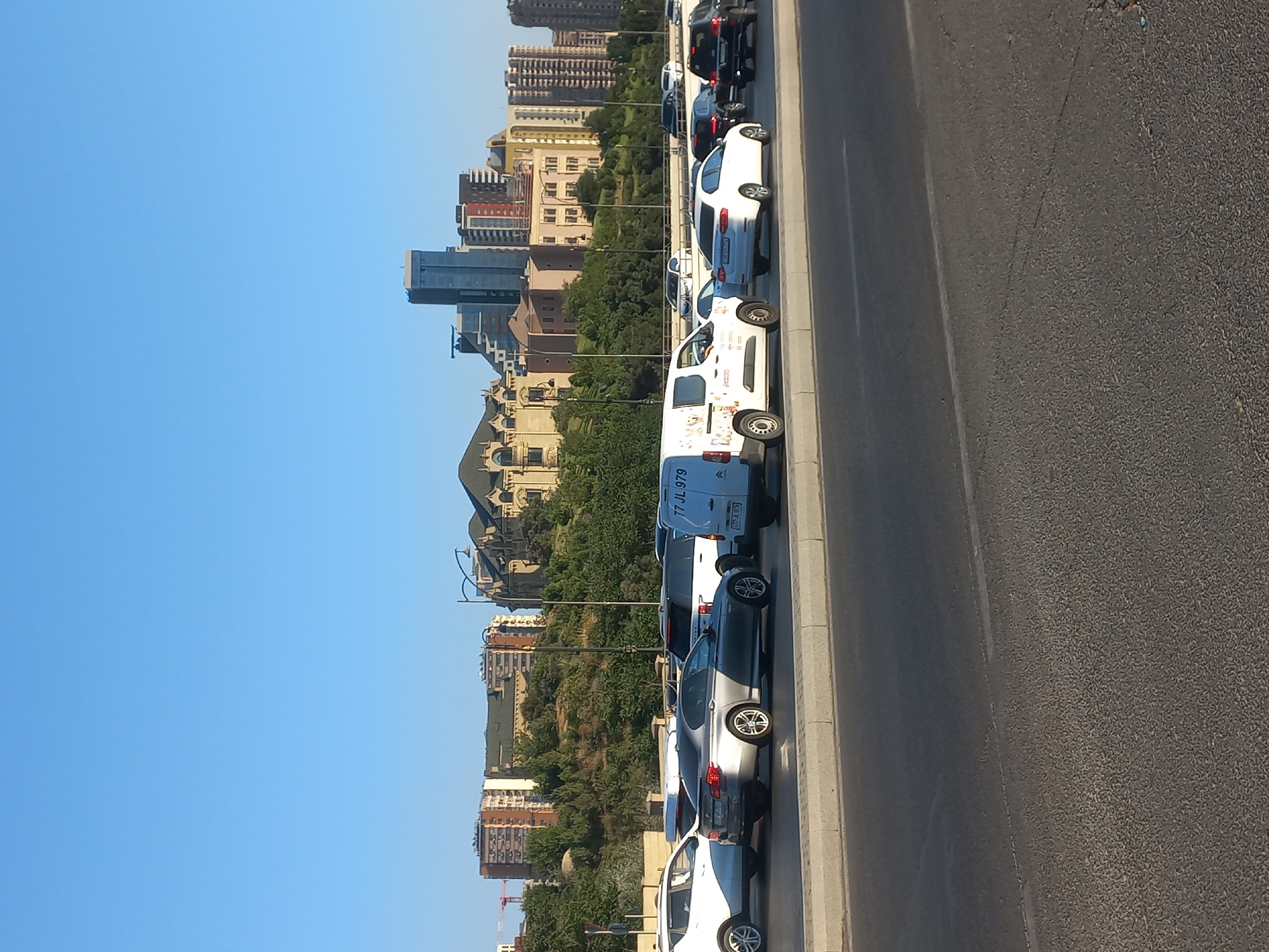 The image depicts a view of the Baku city skyline from a road. In the foreground, several cars are stopped in traffic on a multi-lane road. Vehicles visible include a silver BMW sedan, a white van with a colorful decal, a white SUV, and other passenger cars. The road has a concrete barrier on the left and a concrete curb separating it from the shoulder. In the background, there is a green, tree-covered hill on which various buildings are situated. These buildings include modern high-rise structures, as well as older, ornate architectural styles. The sky above is clear and blue. The overall scene suggests a typical urban environment in Baku, likely during daylight hours.
