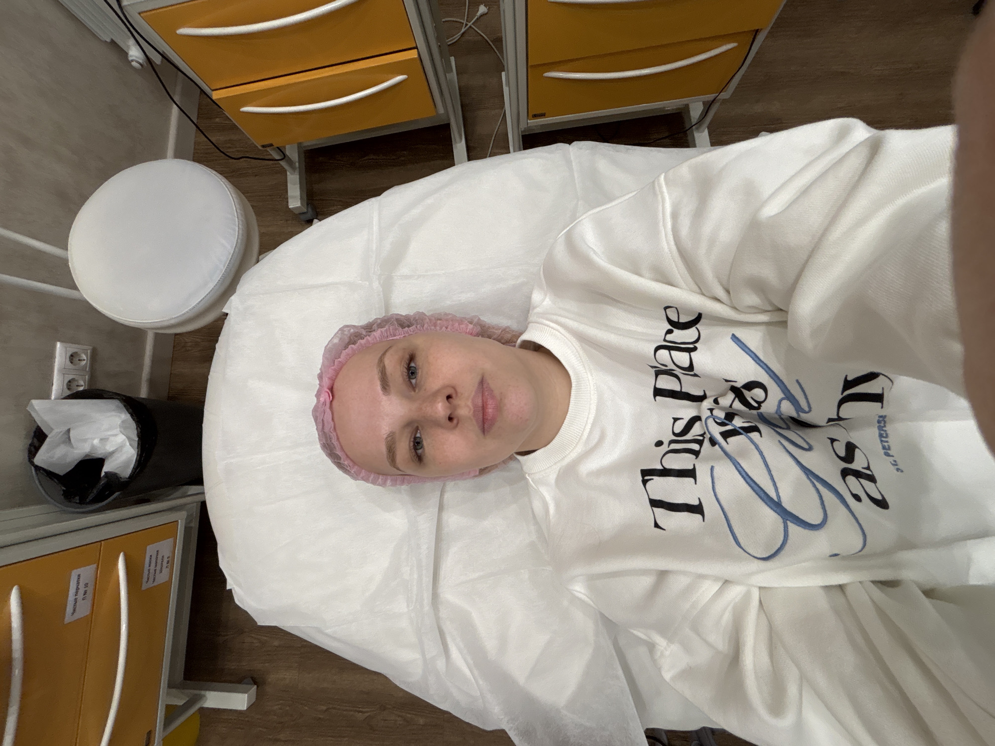 A woman with fair skin, light eyes, and blonde hair covered by a pink surgical cap lies on a white examination table. She wears a white sweatshirt with the text "This Place Your Lash" printed on it in black and blue lettering. The setting is a medical or cosmetic treatment room in Podosenikha, Russia. In the background, there are yellow storage cabinets with white handles, a round white stool, and a trash receptacle. The lighting is bright and artificial. The image appears to be a selfie taken by the woman.