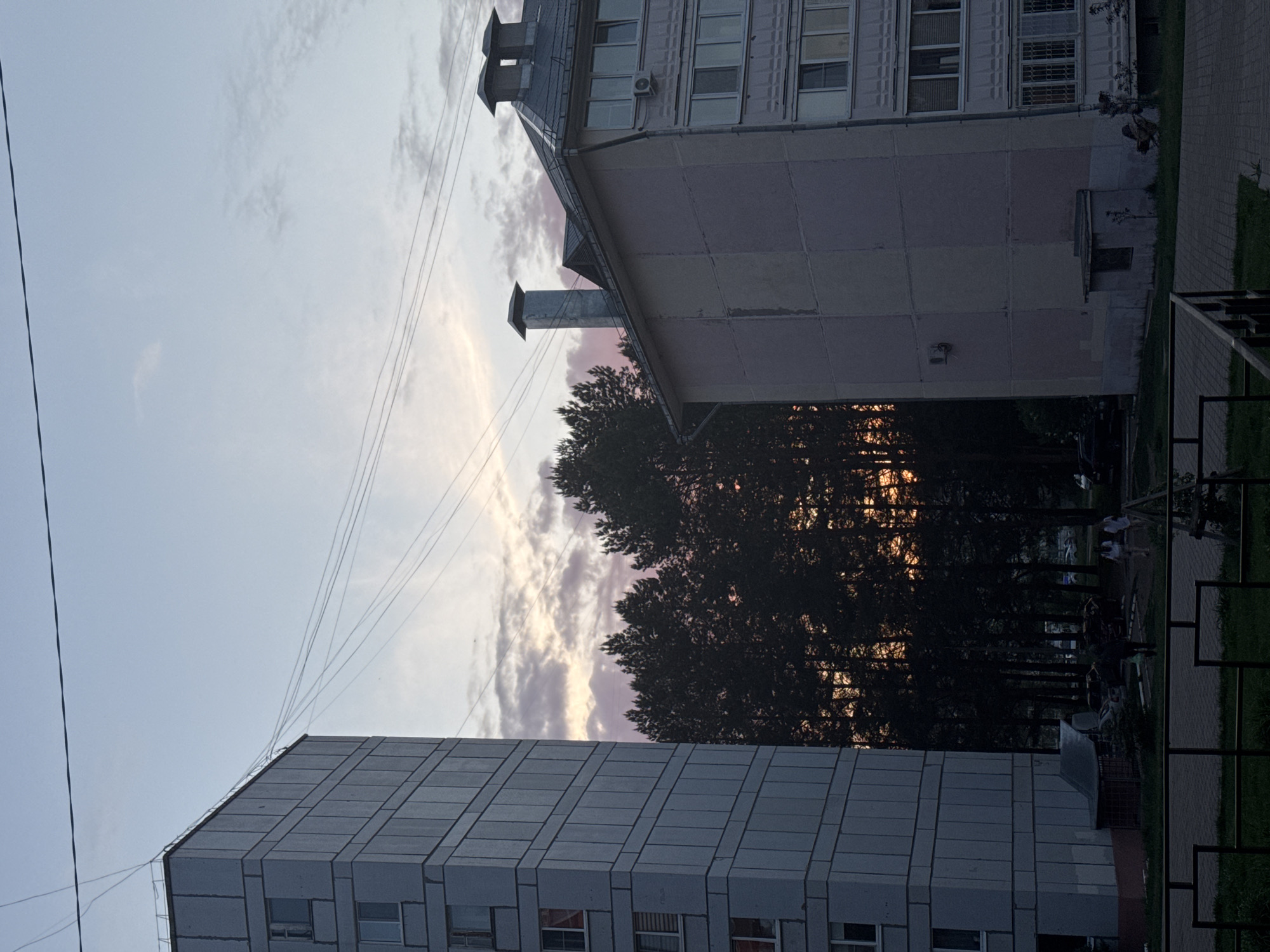 The image shows a view between two buildings in what appears to be an urban area. The building on the left is a tall, grey, multi-story structure with a regular grid pattern of windows. On the right is another building, shorter, with a chimney and what seems to be an air conditioning unit on the wall.
In the middle of the frame, a line of trees obscures the view of a sunset, which is visible between the trees. Power lines run from the top left to the right, connecting the buildings to an electrical grid. Below the trees, there is a park area with people walking around and park structures.
The image is taken in Chaikovsky, Russia.