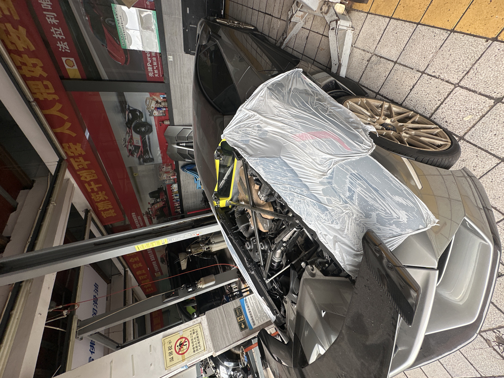 This image depicts the interior of a car repair shop, possibly located in the Chongqing High-tech Zone, China. In the foreground, a dark gray, high-performance sports car, possibly a Lamborghini, is parked with its engine compartment open and partially covered with clear plastic sheeting. The car features a carbon fiber front splitter and gold-colored rims.
The environment suggests a working garage with a car lift positioned behind the sports car. Tools and equipment are visible in the background, along with shelving units that display various automotive products, including bottles of oil. The walls are adorned with large red banners featuring Chinese characters and images of racing cars and oil brands, such as Shell. A yellow warning sign with Chinese text and graphics indicating prohibited entry for unauthorized personnel is affixed to a pillar on the left side of the frame. The floor is tiled with a pattern of beige and yellow squares. The lighting suggests an indoor setting, likely illuminated by overhead fluorescent lights and natural light entering from an unseen source. No people are clearly visible in the frame.