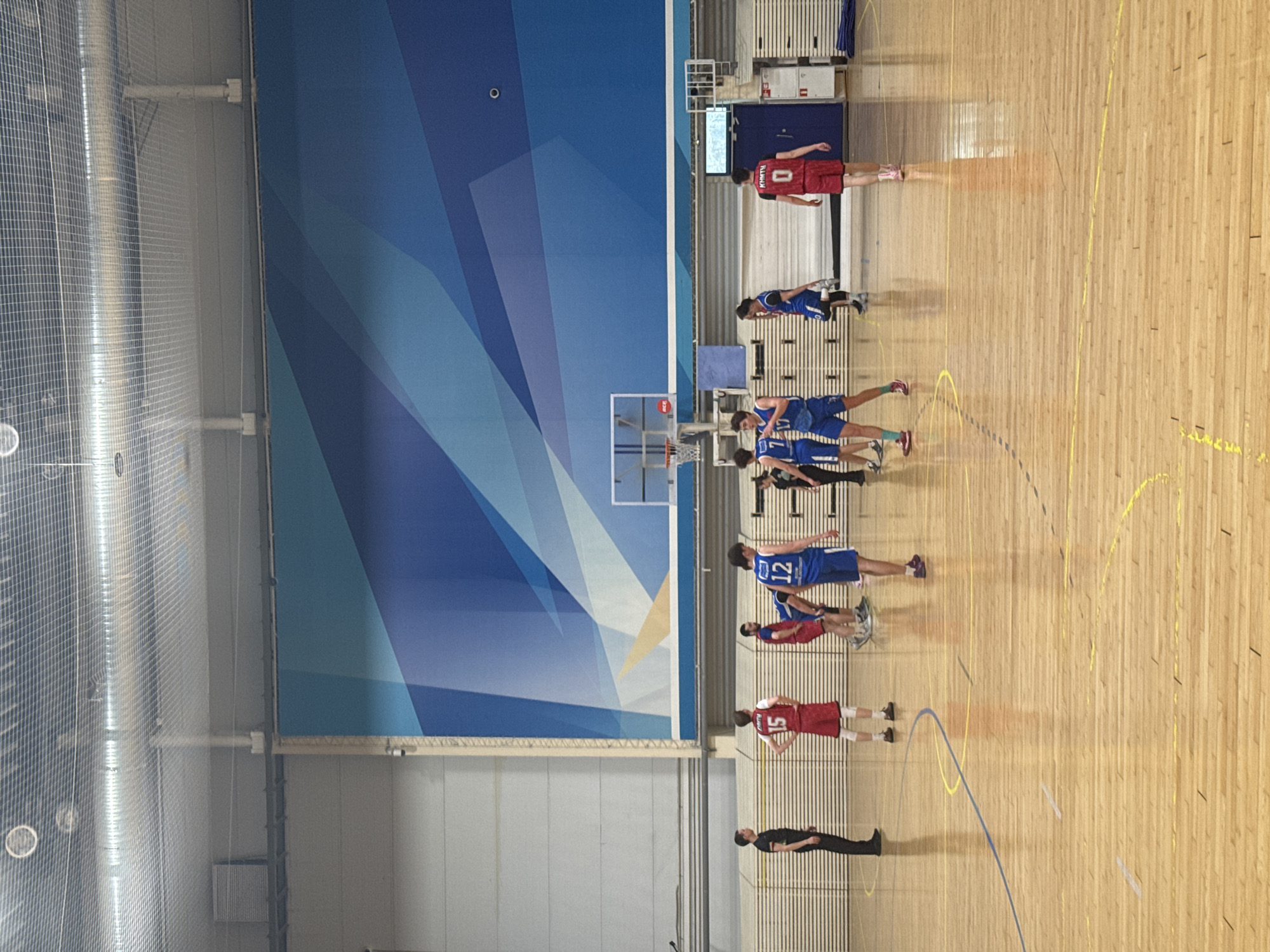 The image shows a basketball game in progress inside a sports hall. One team is wearing red uniforms, and the other is wearing blue. Players are spaced around the free throw lane. A man in a black outfit is standing near the free throw line. The back wall of the court has a large blue graphic. The location is Kazan, Russia.